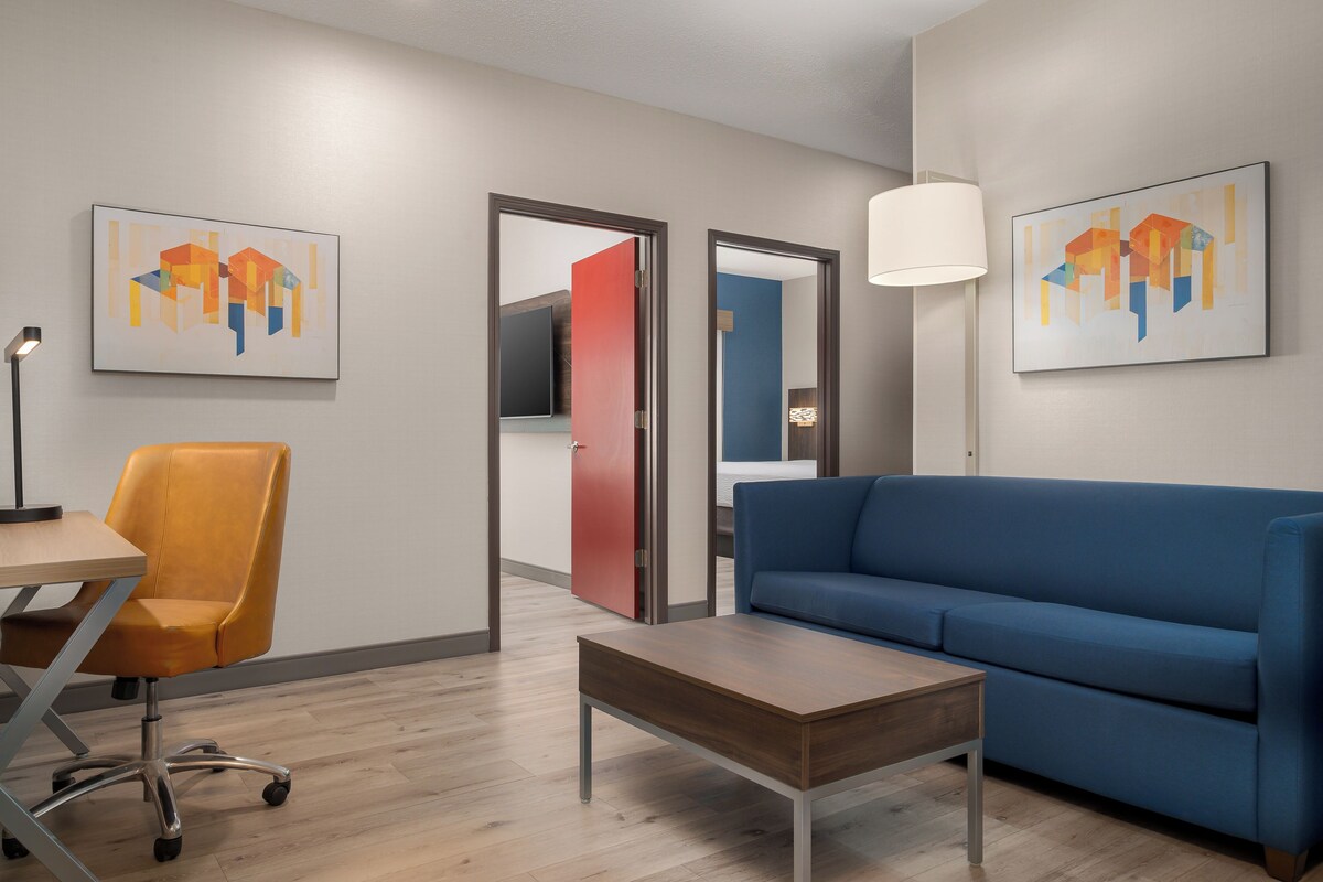 A modern living room with a blue sofa, a wooden coffee table, and a yellow office chair. There are two doors leading to other rooms, and artwork on the walls.