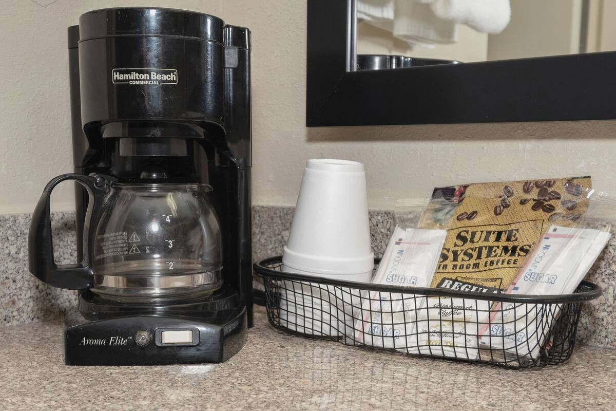 A Hamilton Beach commercial coffee maker, a white coffee cup, and a basket with sugar packets and a coffee bag.