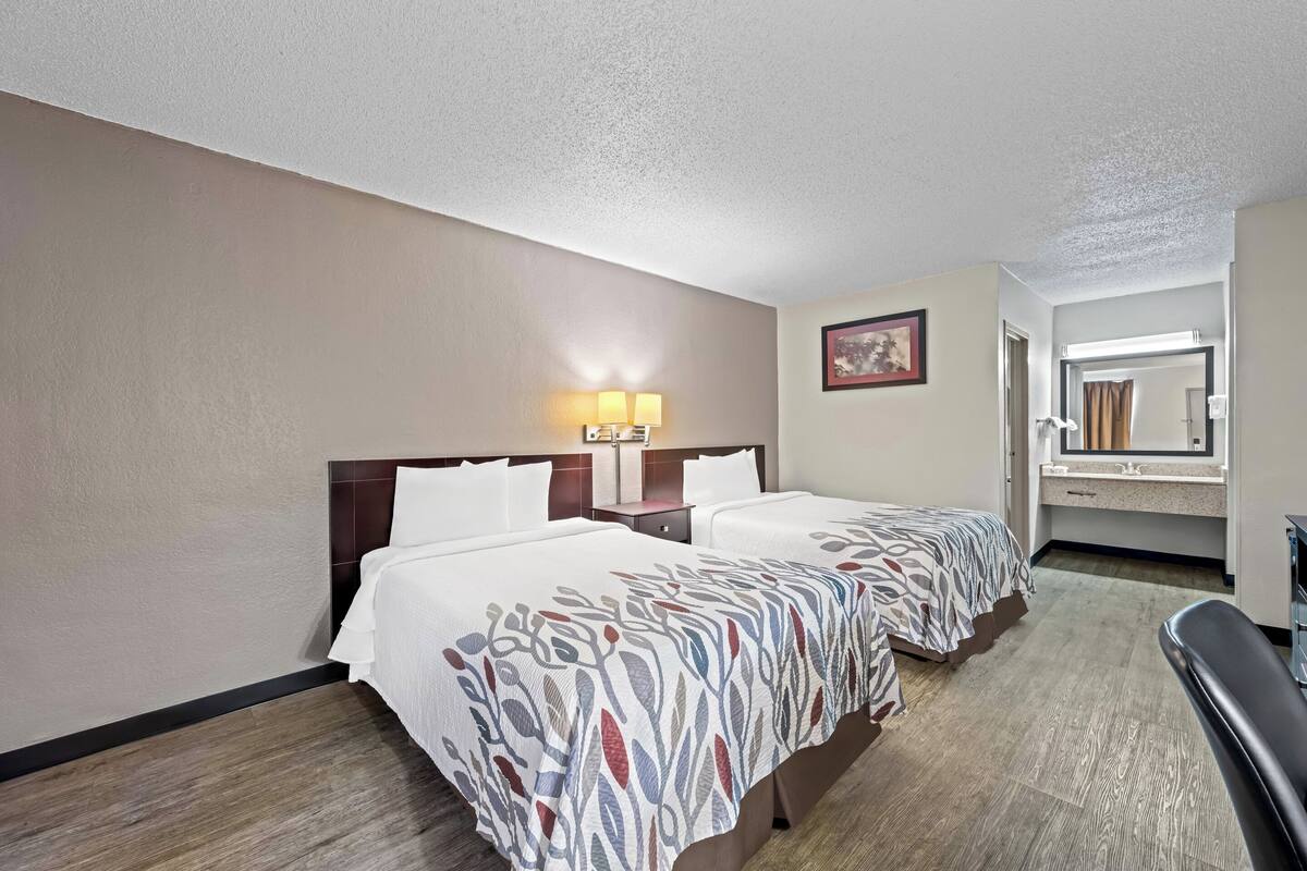 A hotel room with two beds, a nightstand with lamps, a bathroom with a mirror, and a seating area.