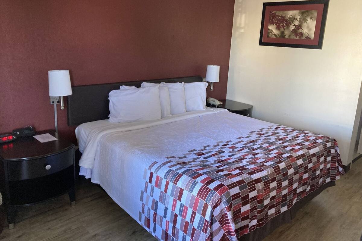 A hotel room with a bed, two bedside tables, a nightstand, a lamp, a clock, and a framed picture on the wall.