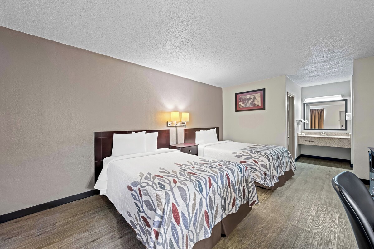 A hotel room with two beds, a nightstand with lamps, a bathroom with a mirror, and a seating area.