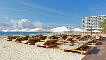 Ein Strand mit Reihen von Holzliegen unter weißen Sonnenschirmen, einem hölzernen Steg und einem Hotelgebäude im Hintergrund.