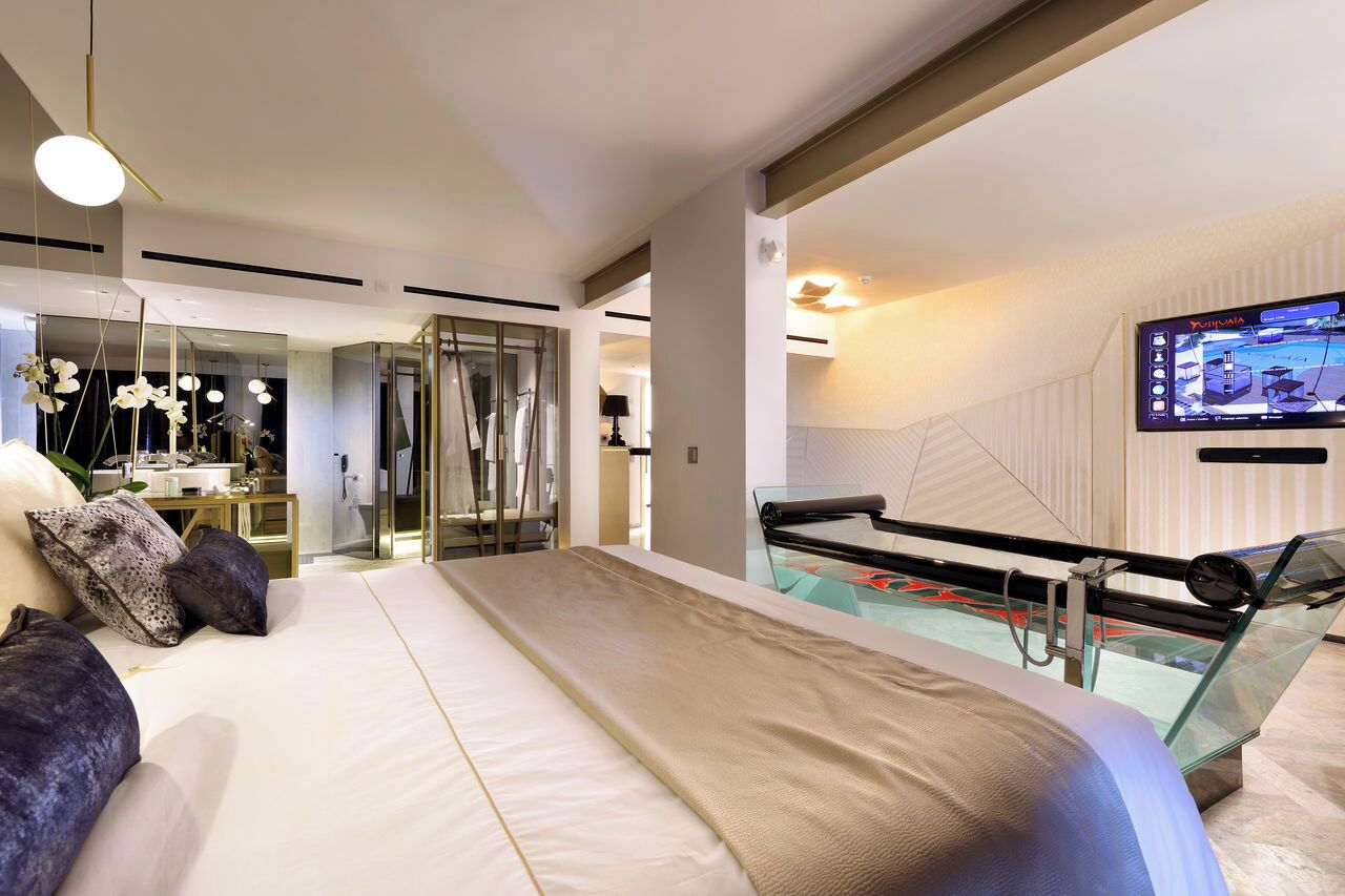 Une chambre d’hôtel moderne avec un grand lit, une table de billard en verre, un téléviseur fixé au mur et un miroir qui reflète la pièce.