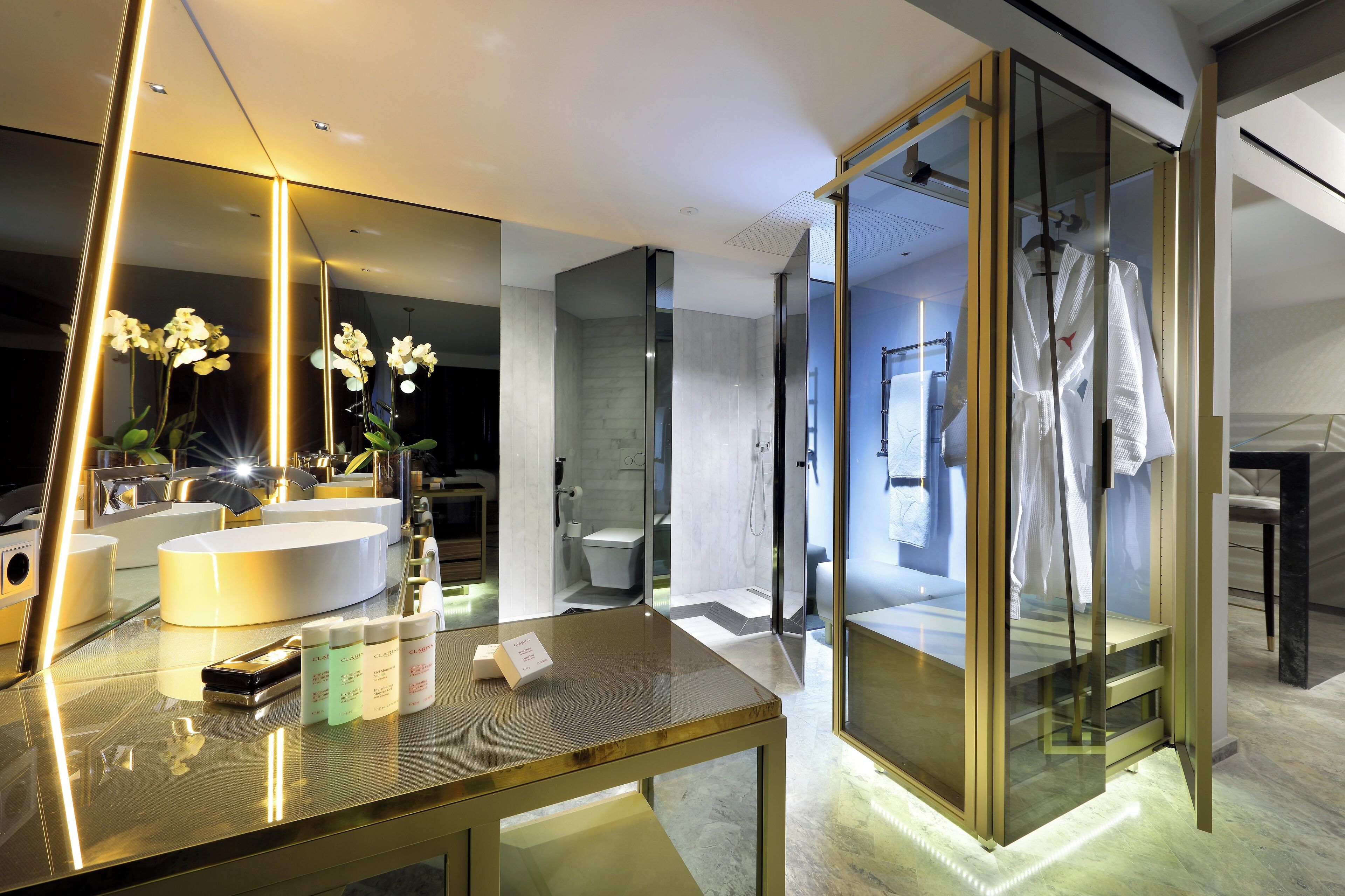 Un baño moderno con cabina de ducha de vidrio, un espejo grande y un lavabo con plantas de orquídea.