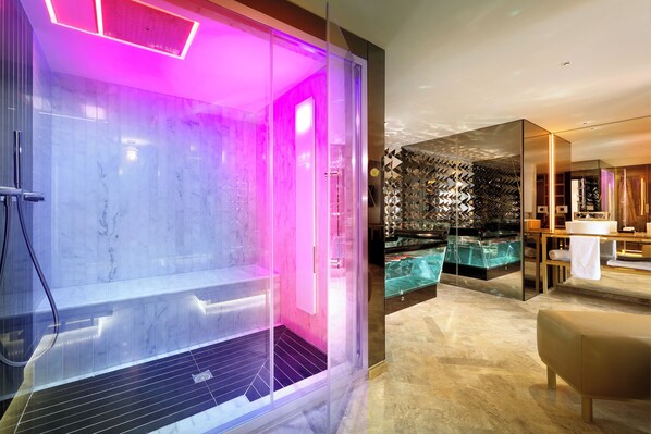 Un moderno espacio de spa con un jacuzzi de paredes de cristal, zona de ducha con azulejos de mármol y un área de descanso con banca de madera.