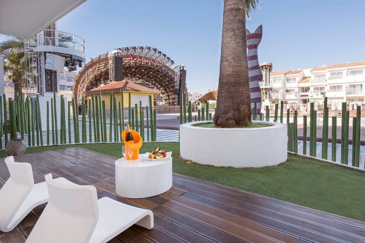 Eine Terrasse mit weißen Liegestühlen, einem kleinen Tisch mit Flasche und Glas sowie einer dekorativen Skulptur.