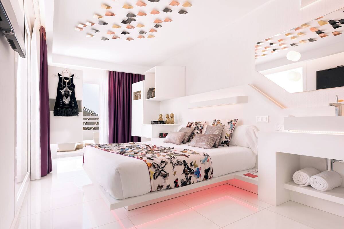 Ein modernes Schlafzimmer mit einem weißen Bett, einem großen Fenster mit violetten Vorhängen, einer Decke mit geometrischen Leuchten und einer weißen Kommode mit Spiegel.