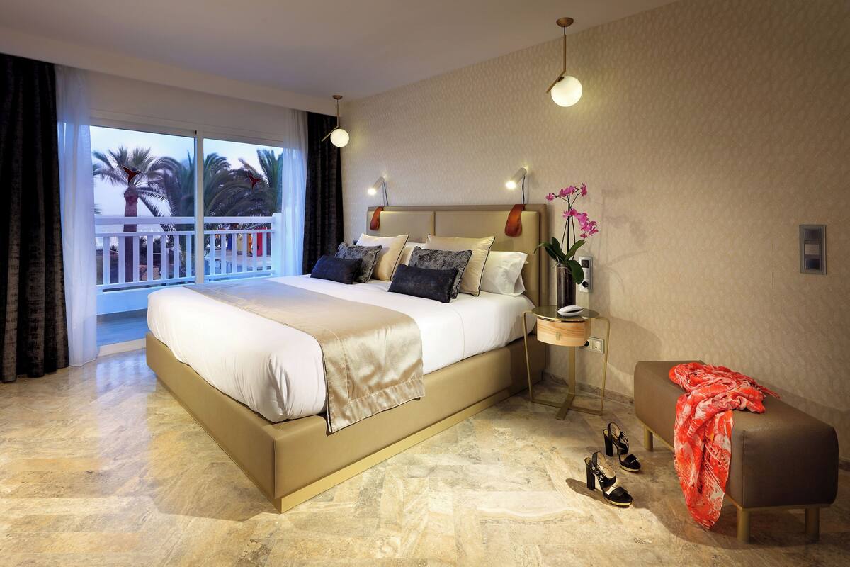 Ein modernes Hotelzimmer mit einem großen Bett, einer kleinen Sitzecke, einem Nachttisch und einem Balkon mit Blick auf Palmen.
