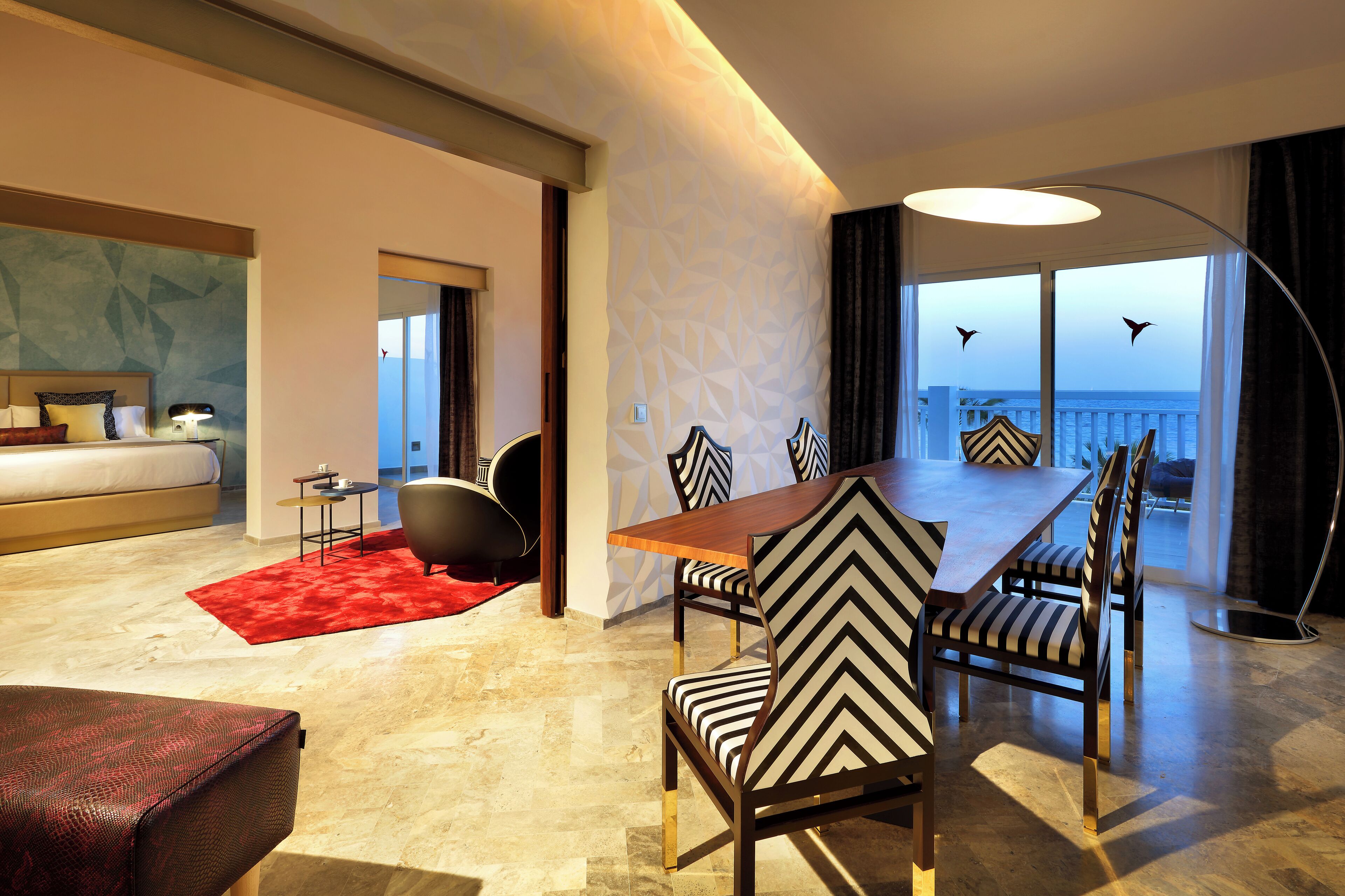 Una habitación de hotel moderna con una cama grande, un comedor con sillas a rayas, una alfombra roja y vistas al mar.