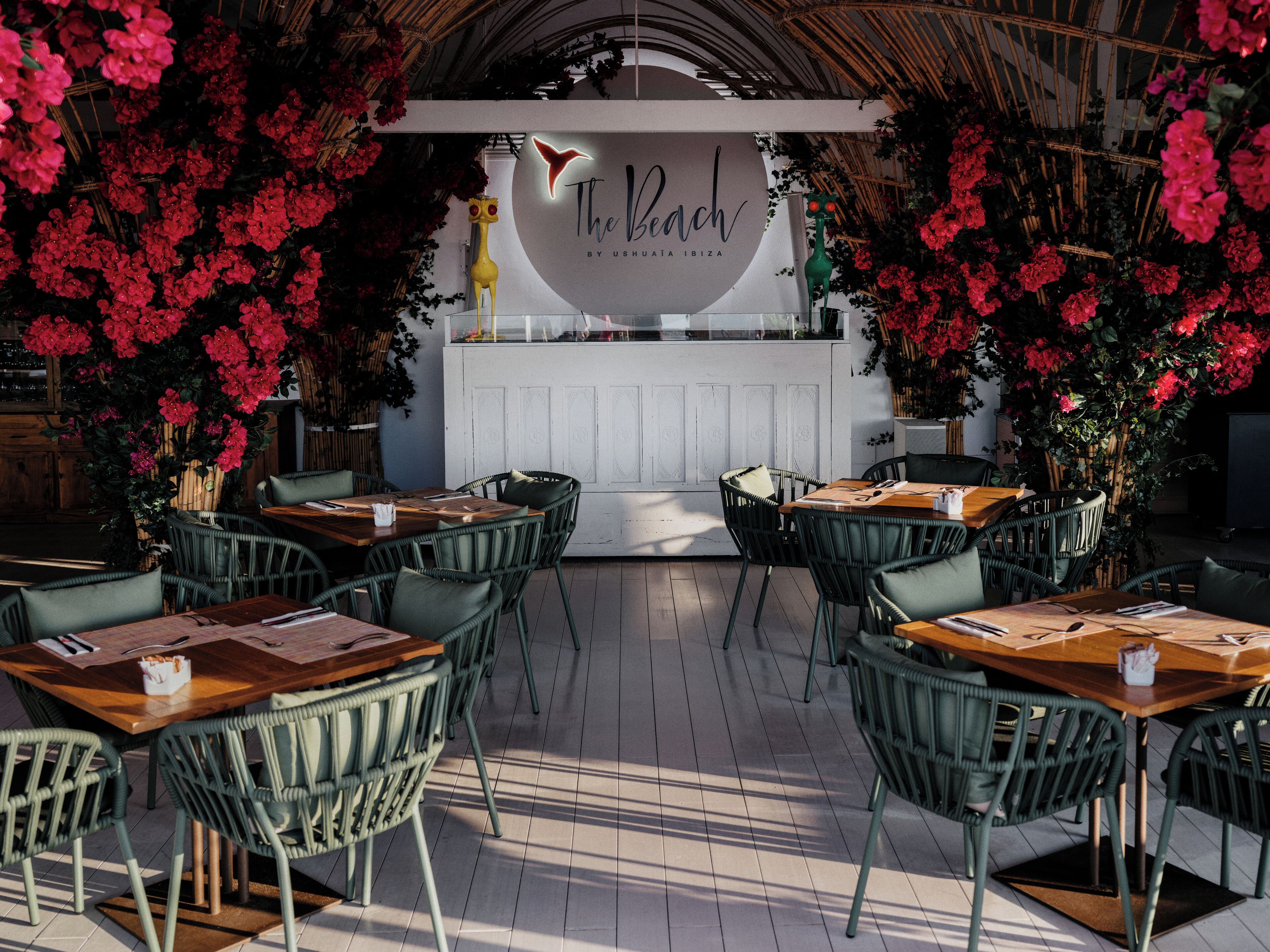 Un espace de restauration en plein air, avec des chaises et des tables en osier, entouré de fleurs rouges et agrémenté d'un comptoir blanc orné d'une enseigne au néon.
