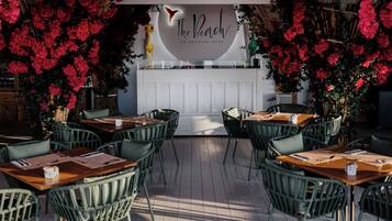 Un espace de restauration en plein air, avec des chaises et des tables en osier, entouré de fleurs rouges et agrémenté d'un comptoir blanc orné d'une enseigne au néon.