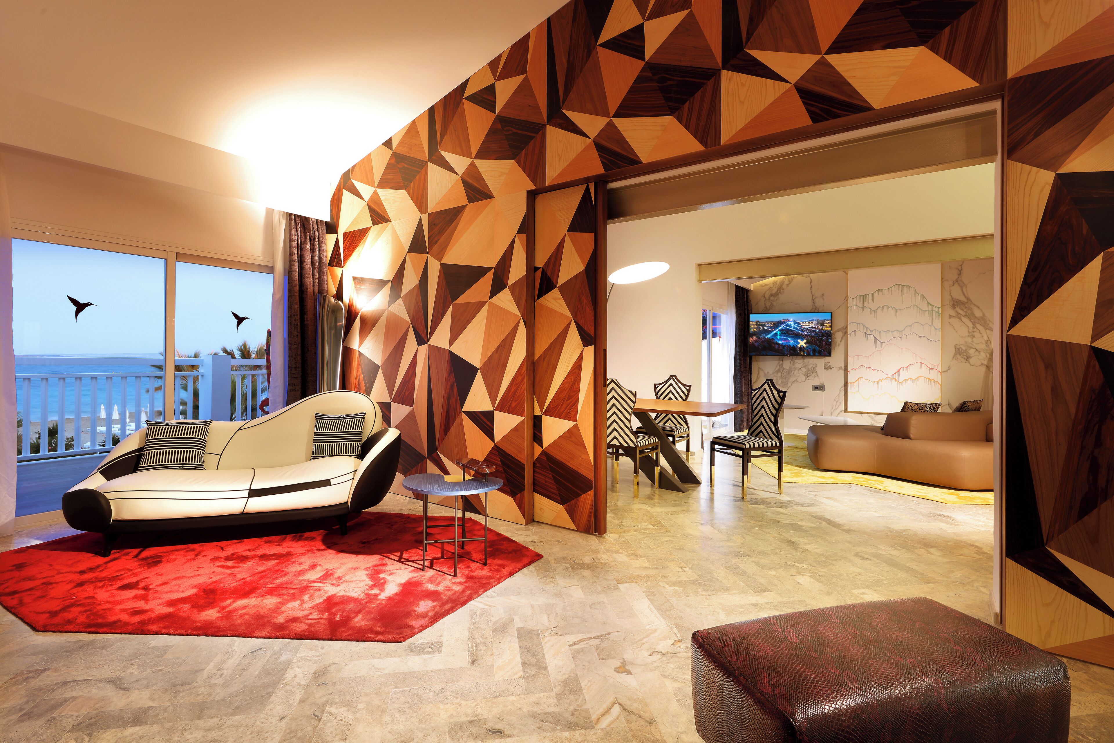 Un salón moderno con una pared de estampado geométrico, un sofá, una alfombra roja y vistas al mar.