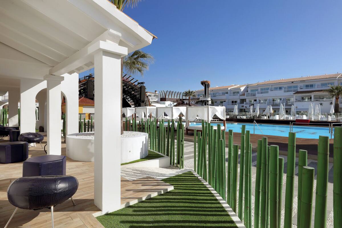 Ein Poolbereich mit weißer Pergola, grünen zylindrischen Absperrungen und einem Pool mit Liegestühlen und Sonnenschirmen.