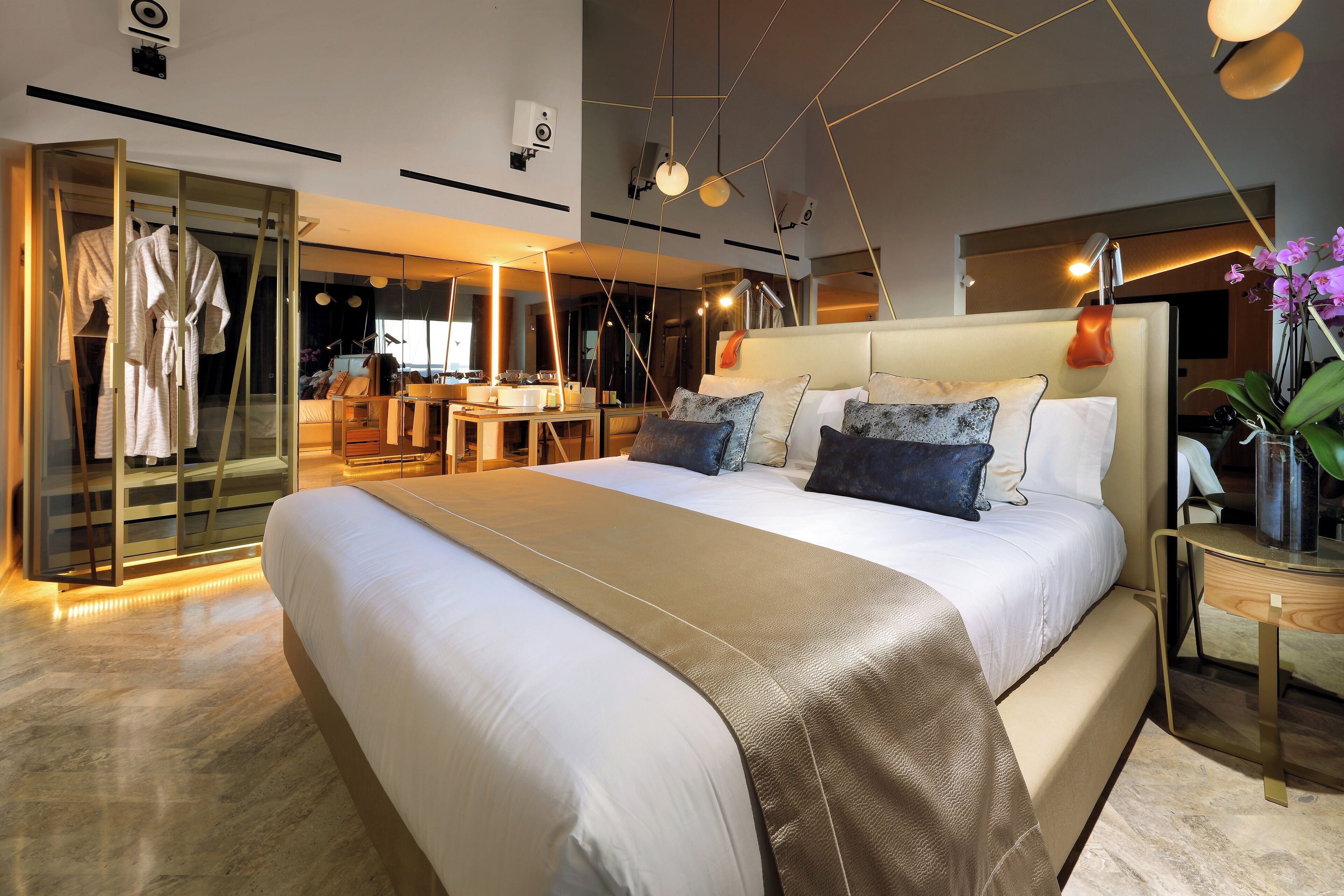 A modern hotel room with a large bed, a glass wardrobe, a desk, and a nightstand with a lamp.