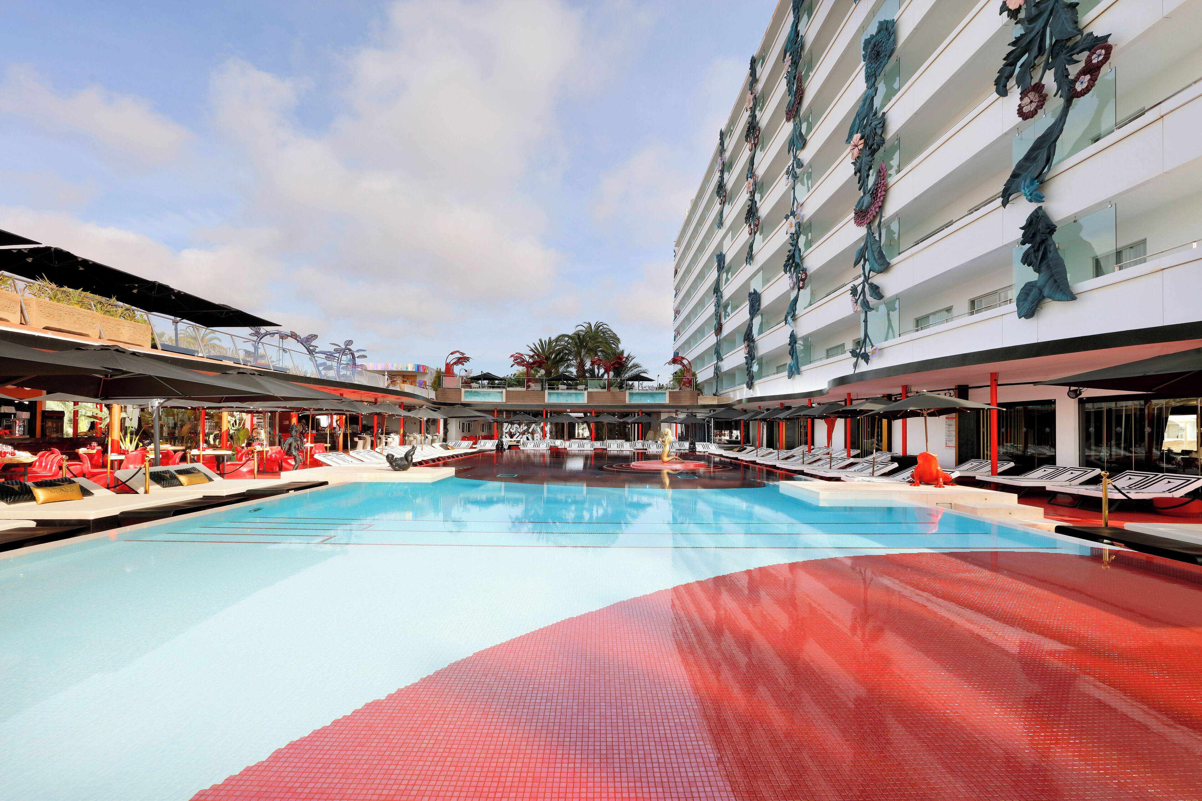 Area piscina di un hotel con diversi livelli di balconi, pavimento in piastrelle rosse e blu, e poltrone da relax rosse.