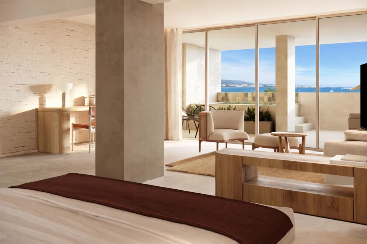 A modern hotel room with a large bed, a seating area with a coffee table, a TV, and a balcony with a view of the sea.