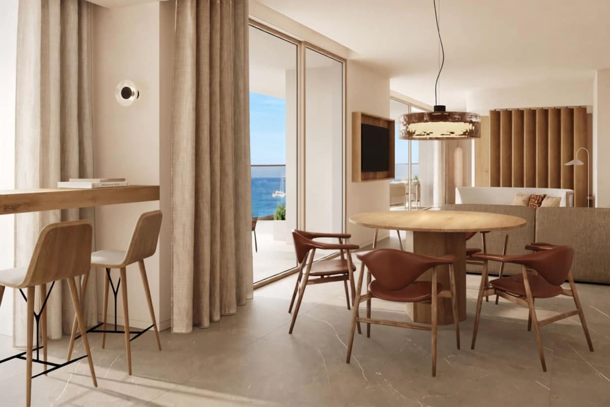 A modern living room with a round wooden table, multiple chairs, a sofa, and a large window offering a view of the sea.