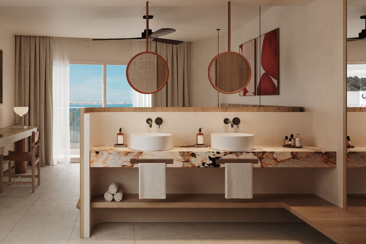 A modern bathroom with two sinks, a ceiling fan, and a view of the sea.