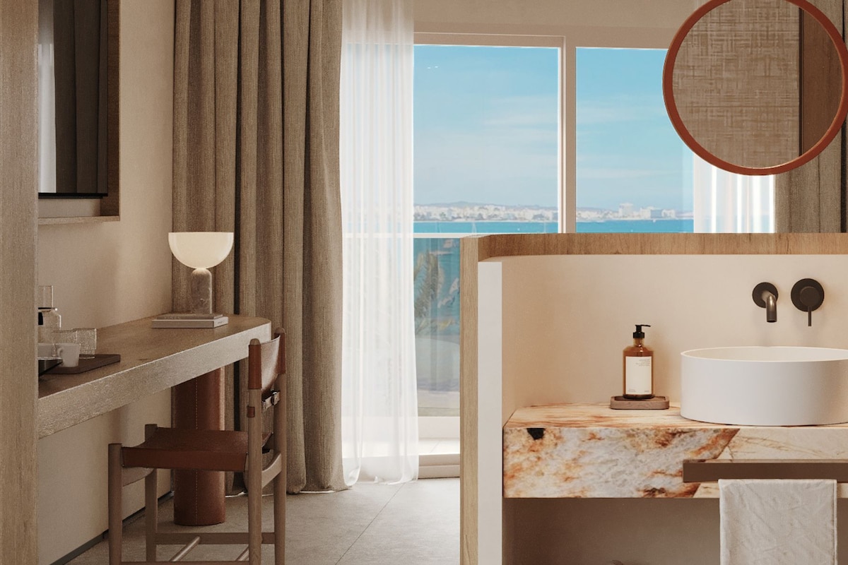 A modern bathroom with a marble countertop, a round mirror, and a view of the city through large windows.