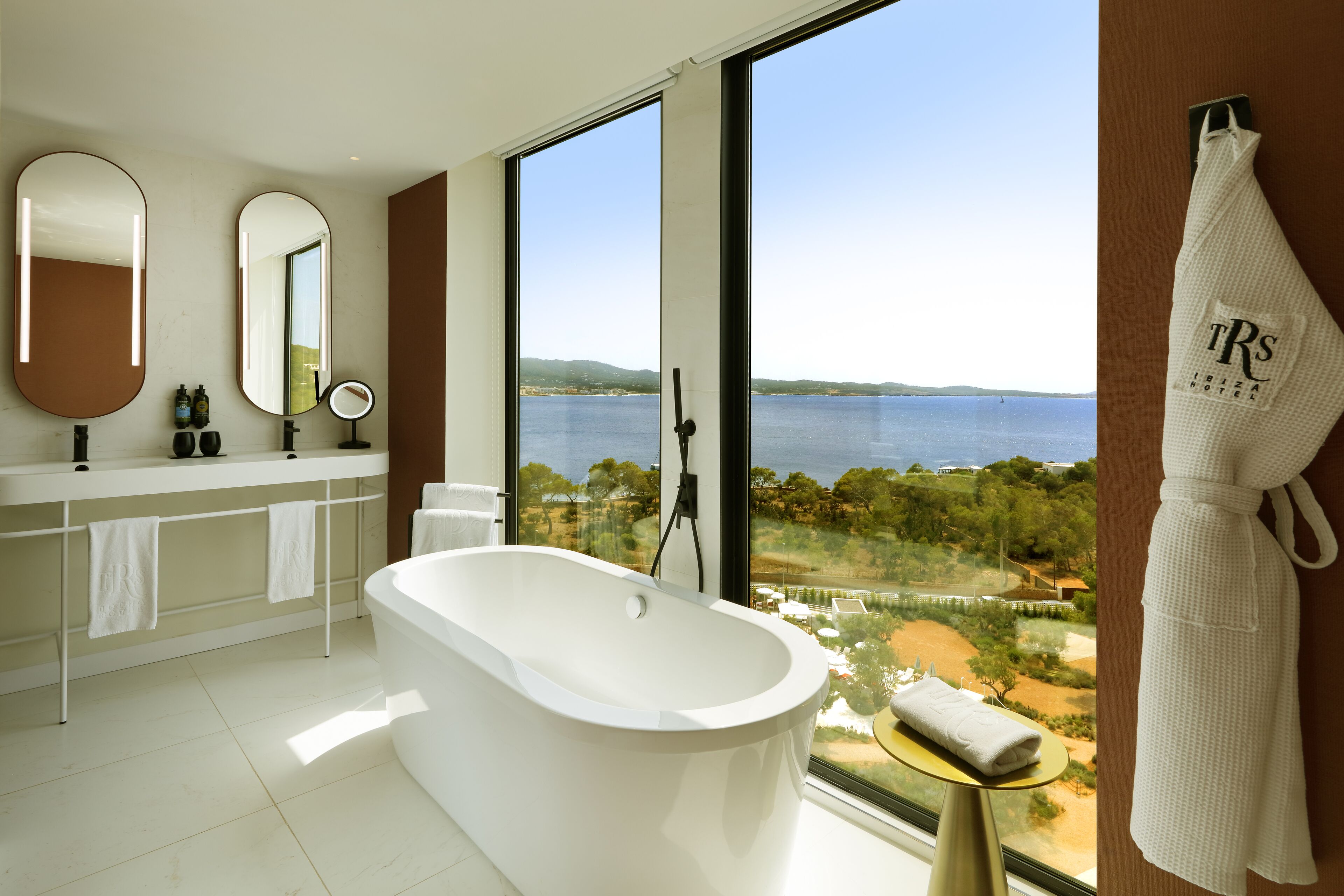 A modern bathroom with a large bathtub, a vanity with a mirror, and a view of the sea.