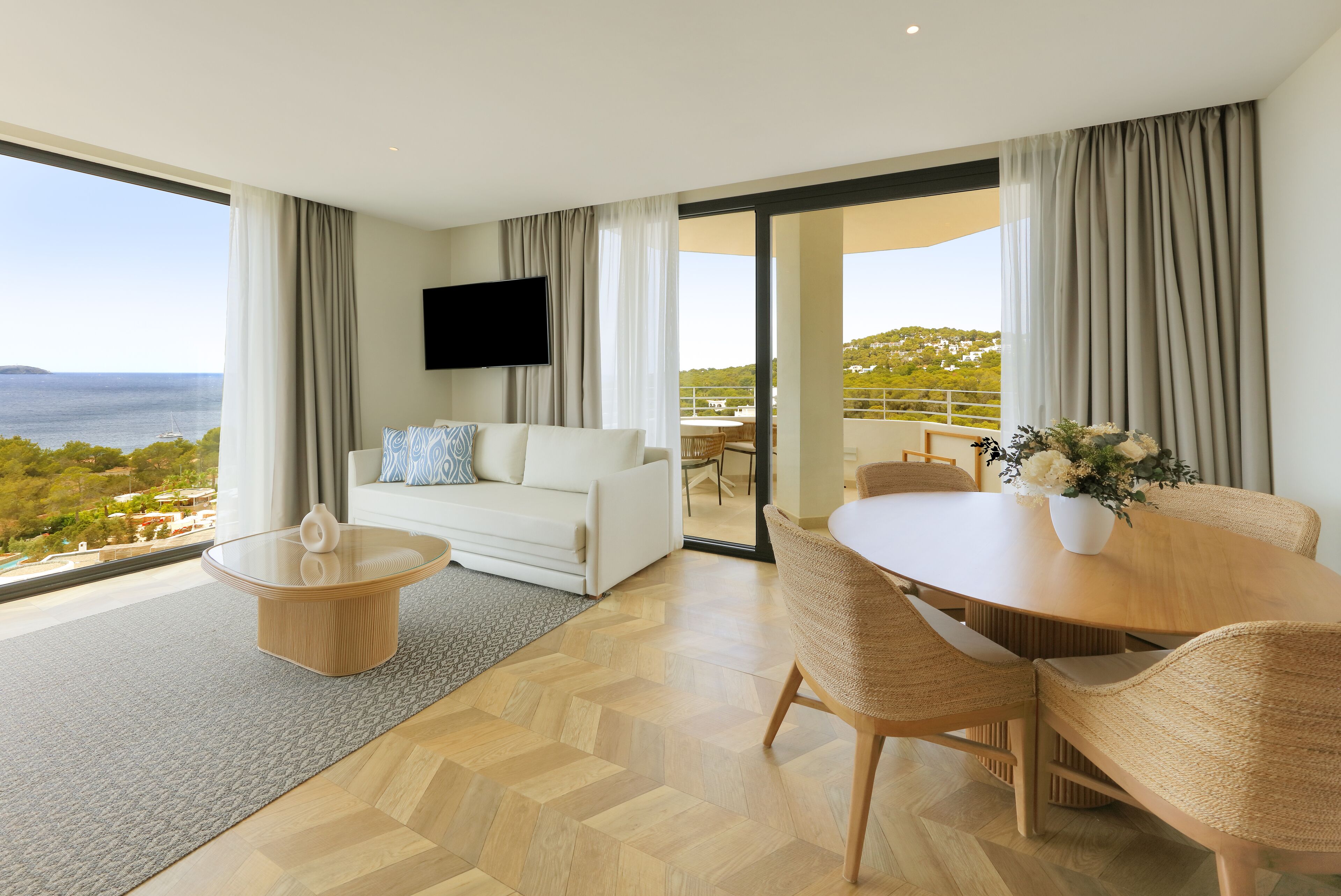 A modern hotel room with a large balcony, a white sofa, a dining table with chairs, and a flat-screen TV.