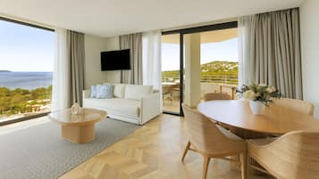 A modern hotel room with a large balcony, a white sofa, a dining table with chairs, and a flat-screen TV.