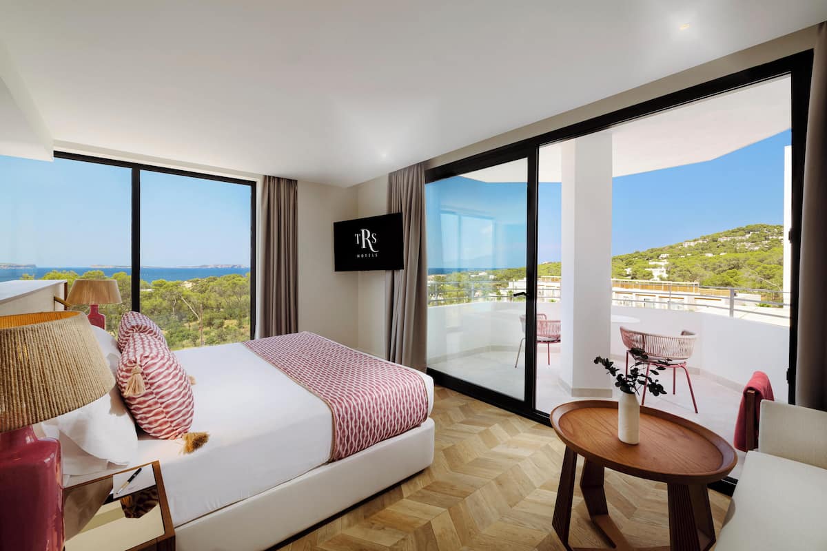 A modern hotel room with a large bed, a TV, a balcony with a view of the sea, and a small table with a vase.