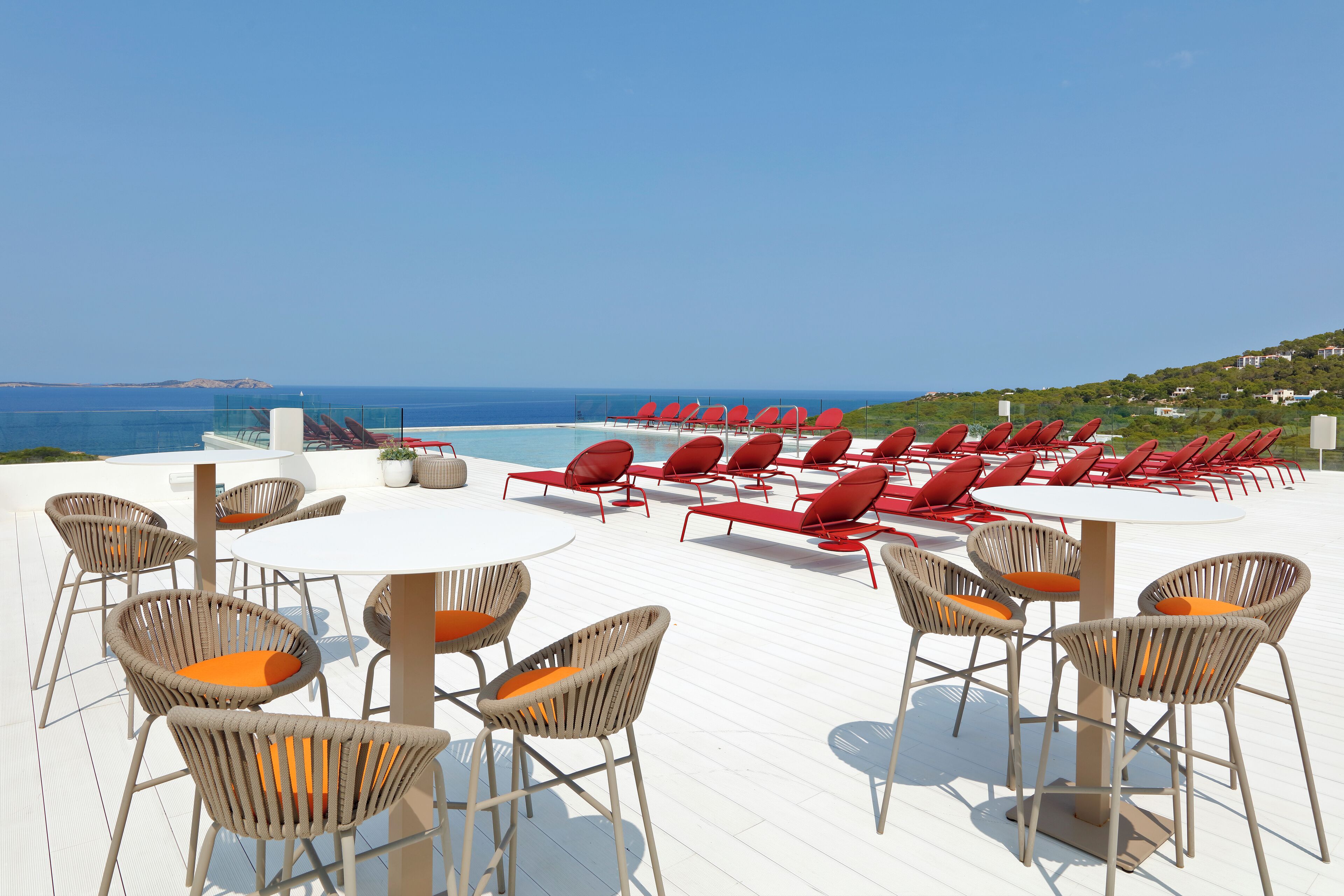 Terraza soleada con sillas de mimbre y una mesa redonda, con vistas a una piscina y al mar.