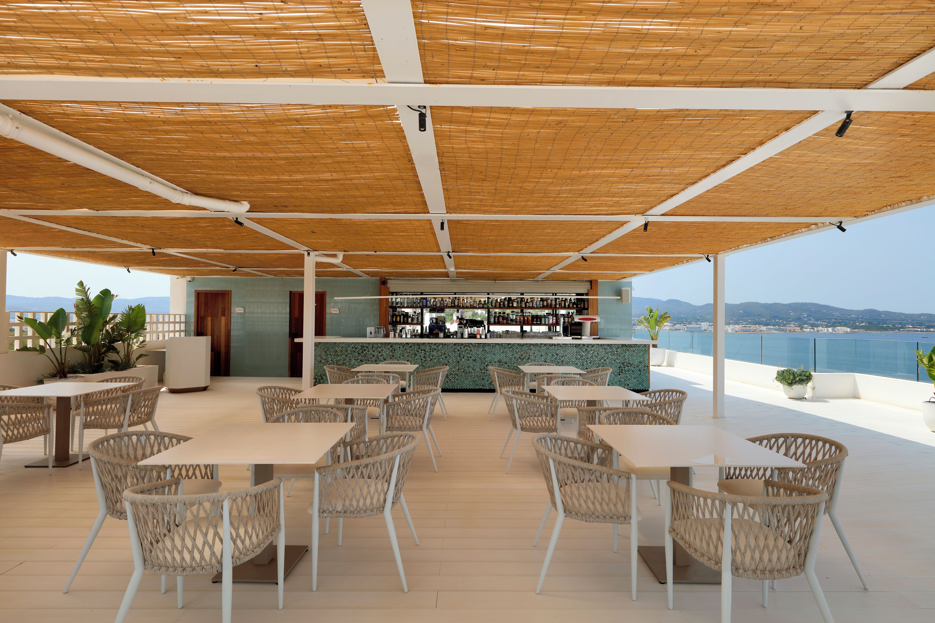 Een terras buiten met tafels en stoelen, een bar achterin en uitzicht op zee.