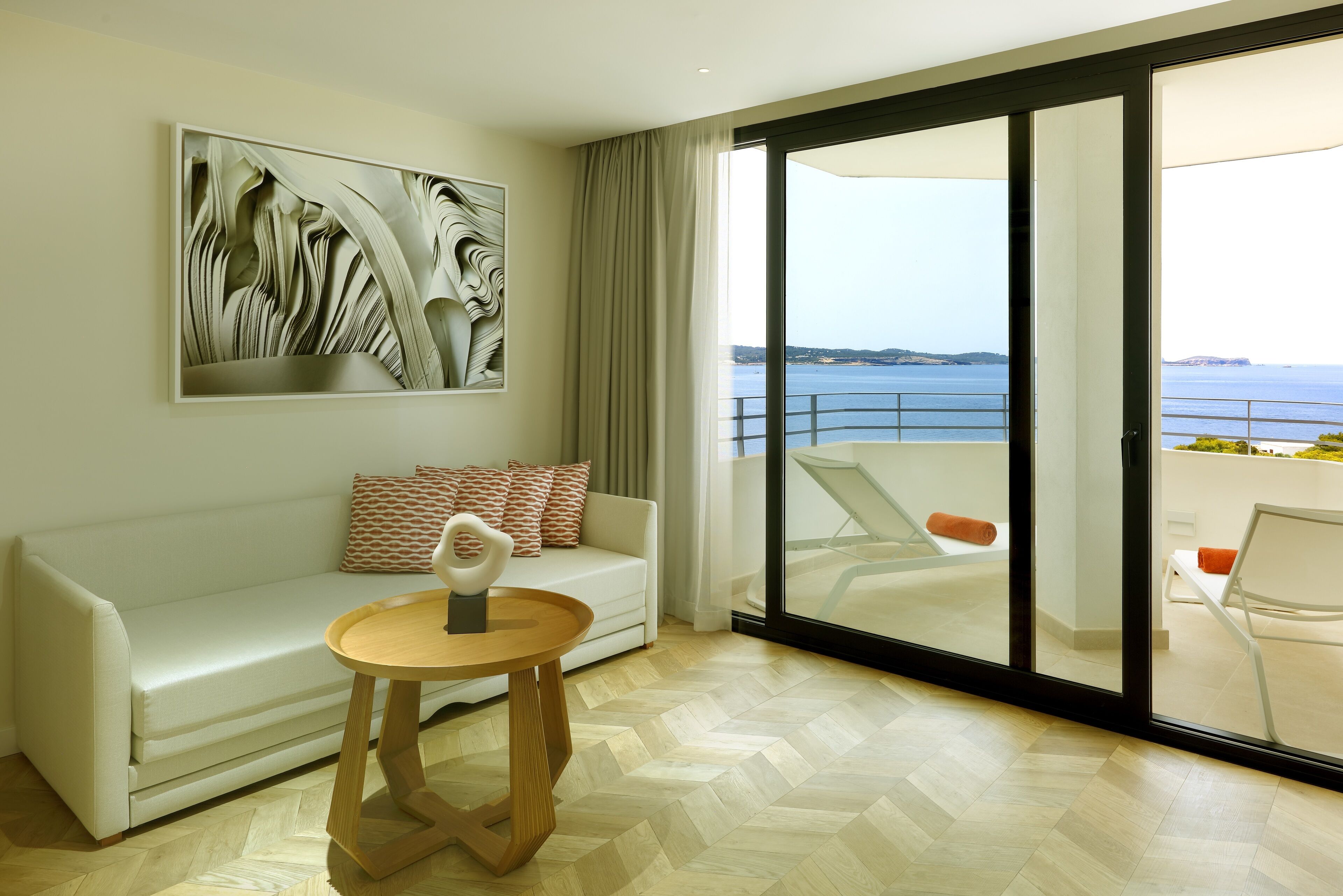 Un salon moderne avec un canapé blanc, une table d’appoint en bois et un grand tableau abstrait. Une porte-fenêtre coulissante donne accès à un balcon avec vue sur la mer.