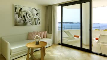 Un salon moderne avec un canapé blanc, une table d’appoint en bois et un grand tableau abstrait. Une porte-fenêtre coulissante donne accès à un balcon avec vue sur la mer.