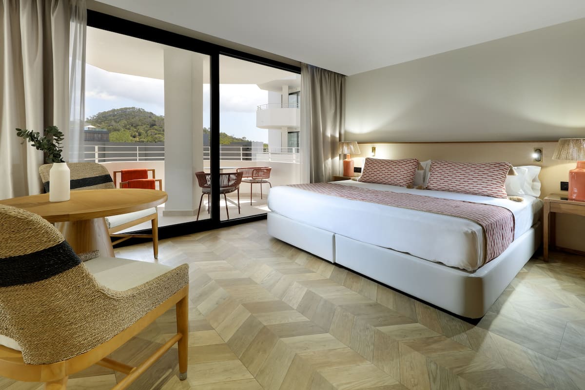 A modern hotel room with a large bed, a dining area with a table and chairs, and a balcony with a view of a hill.