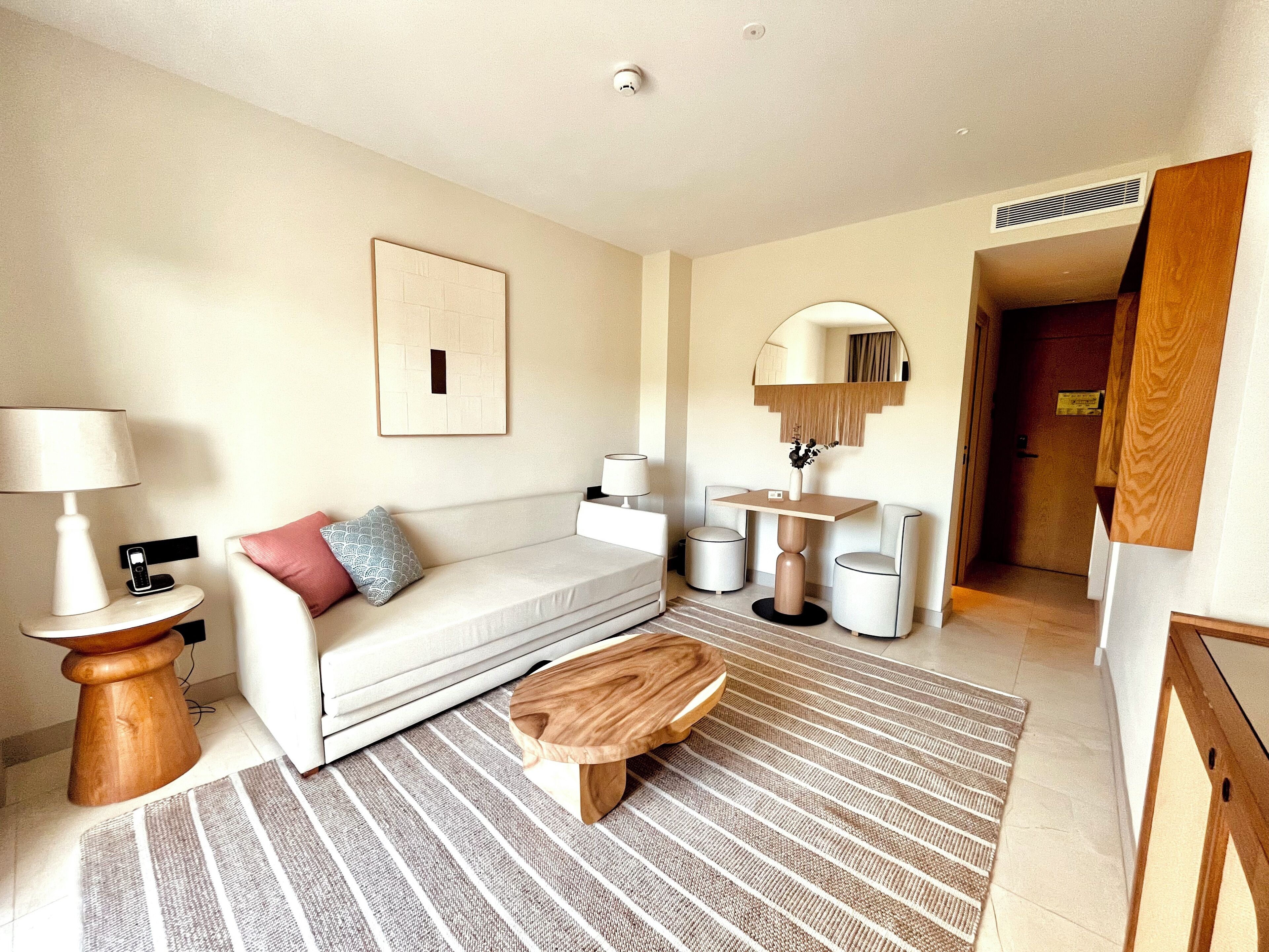 A modern hotel room with a sofa, a wooden coffee table, a lamp, a side table, and a mirror.