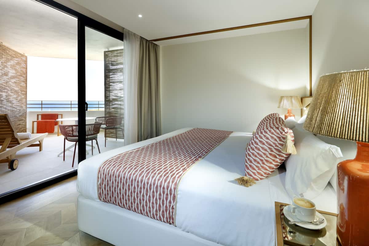 A modern hotel room with a large bed, a dining area with a table and chairs, and a view of the sea through a glass door.