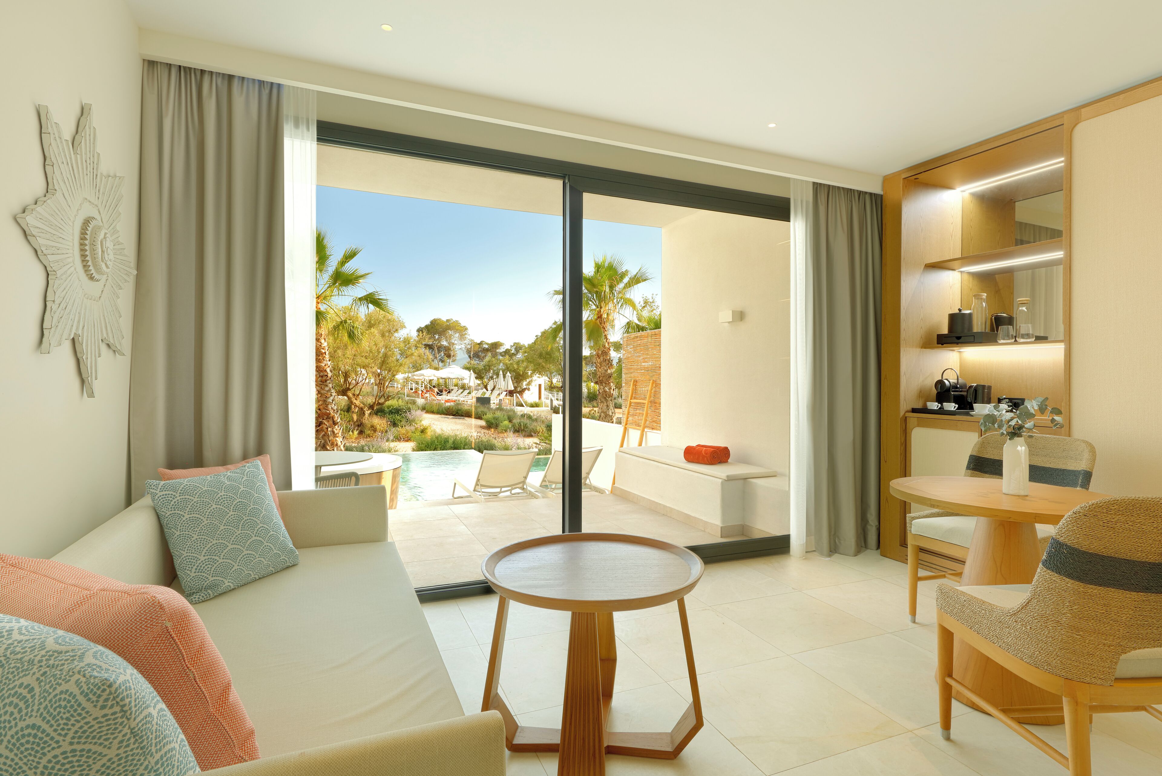 A modern hotel room with a large sliding glass door leading to a pool area, a round wooden table, and a cozy seating area with a sofa and chairs.