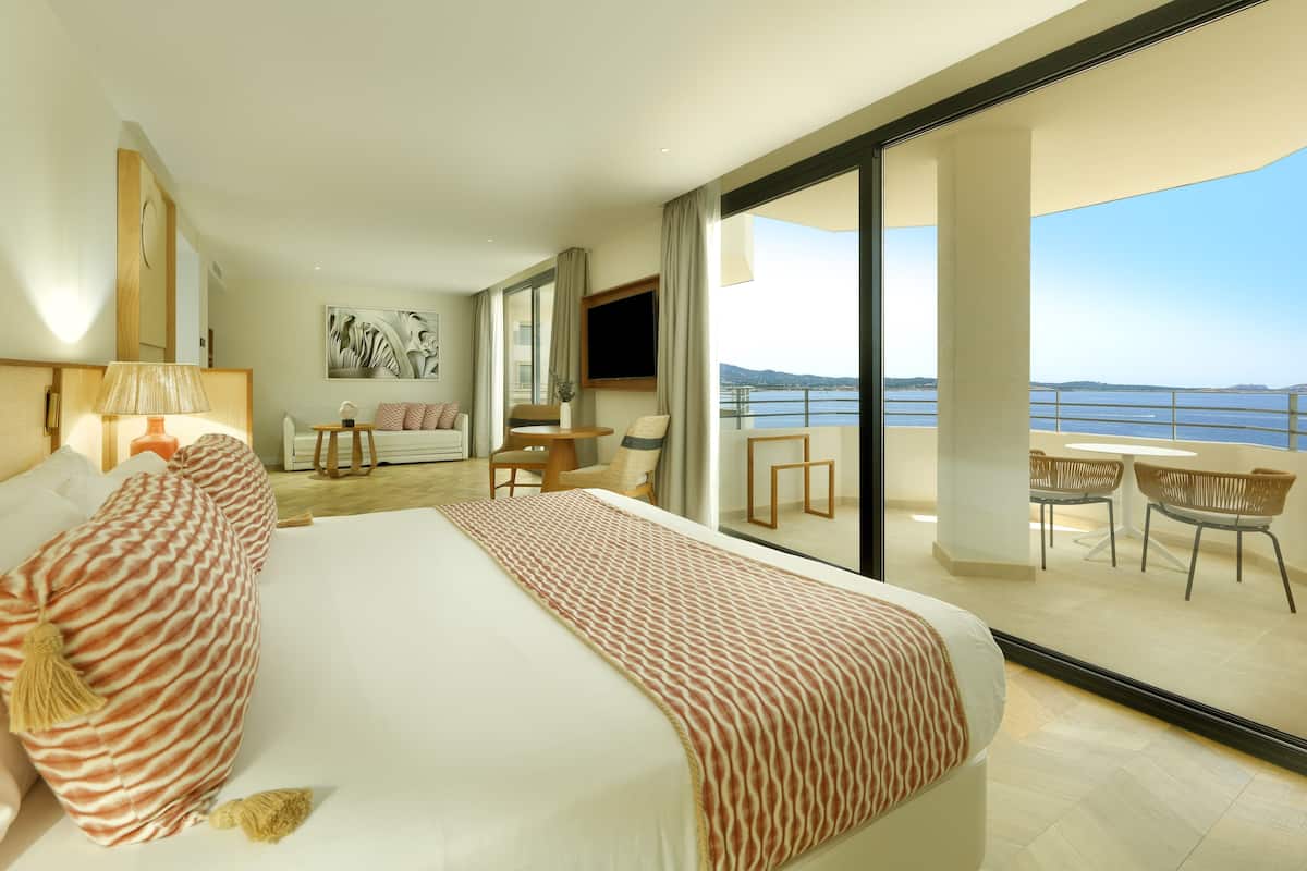 A modern hotel room with a large bed, a TV, a balcony with outdoor seating, and a view of the sea.