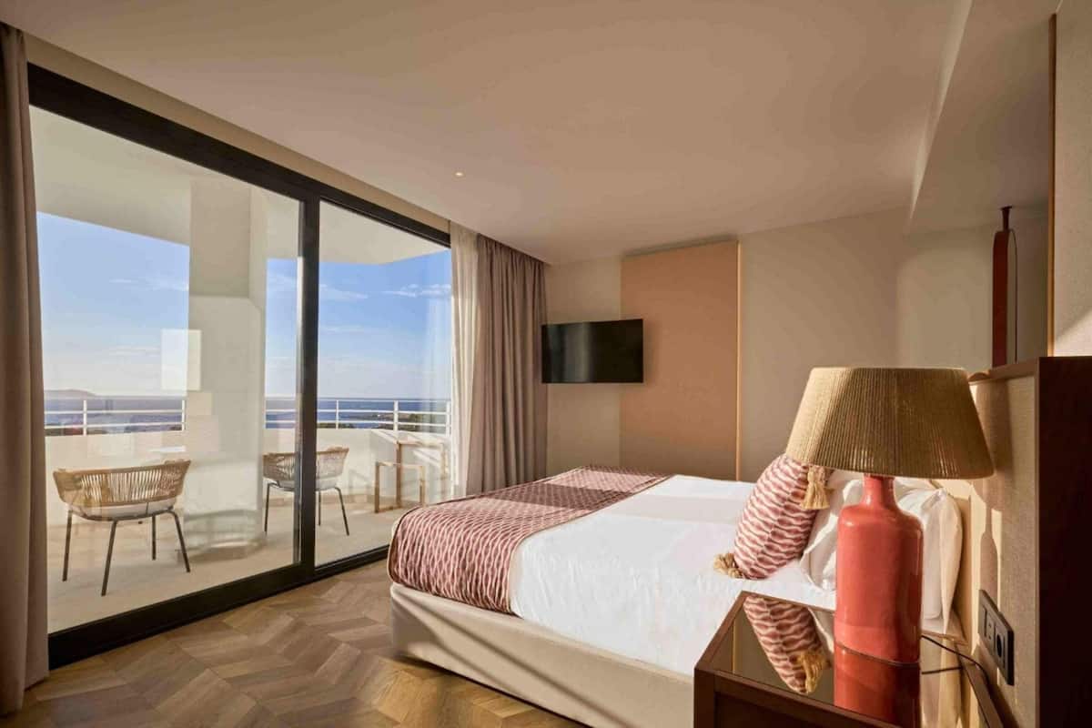 A hotel room with a large bed, a TV, a lamp, and a view of the balcony with chairs.