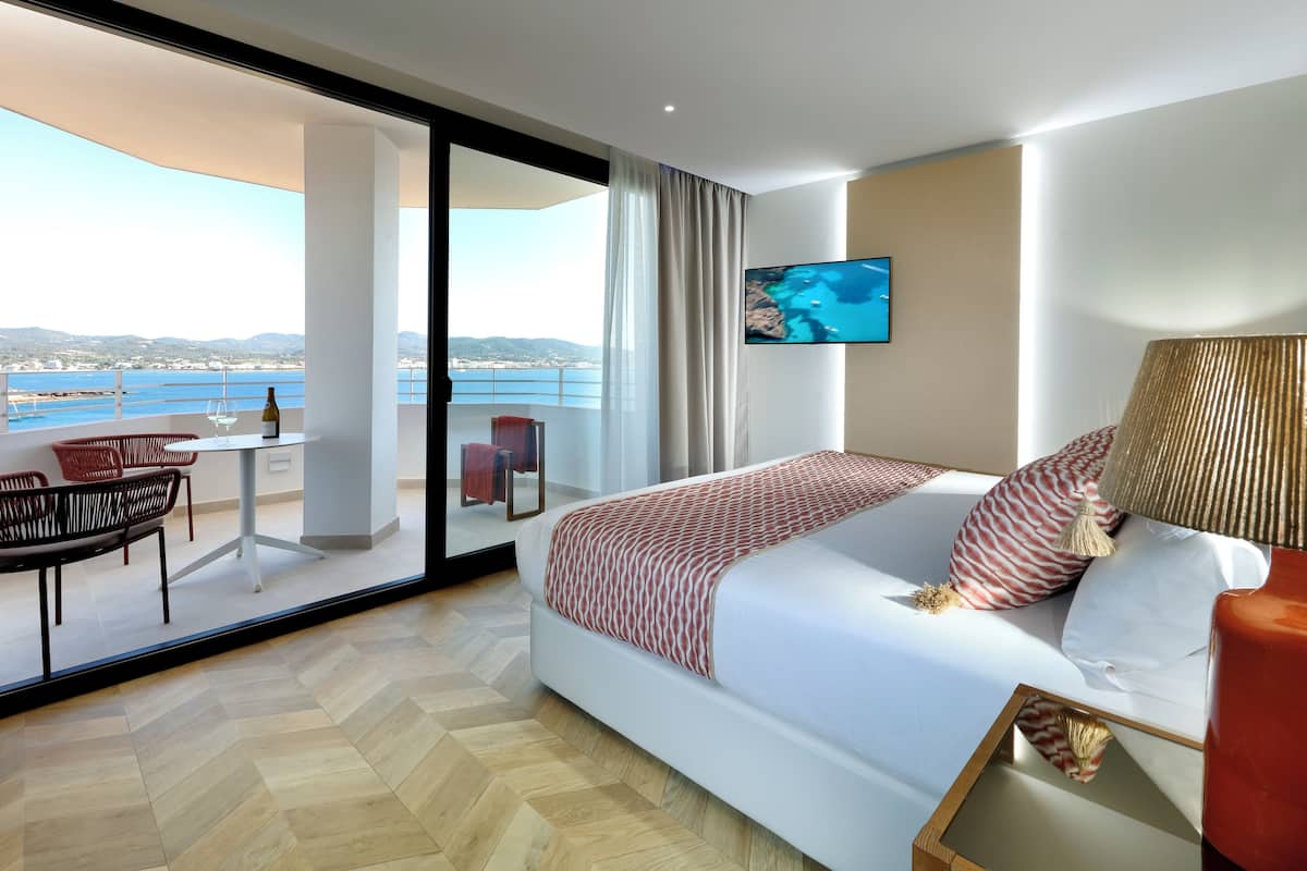 A modern hotel room with a large bed, a TV mounted on the wall, and a balcony with a view of the sea.