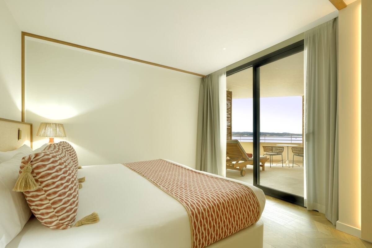 A hotel room with a large bed, a view of the balcony, and a lamp on the nightstand.