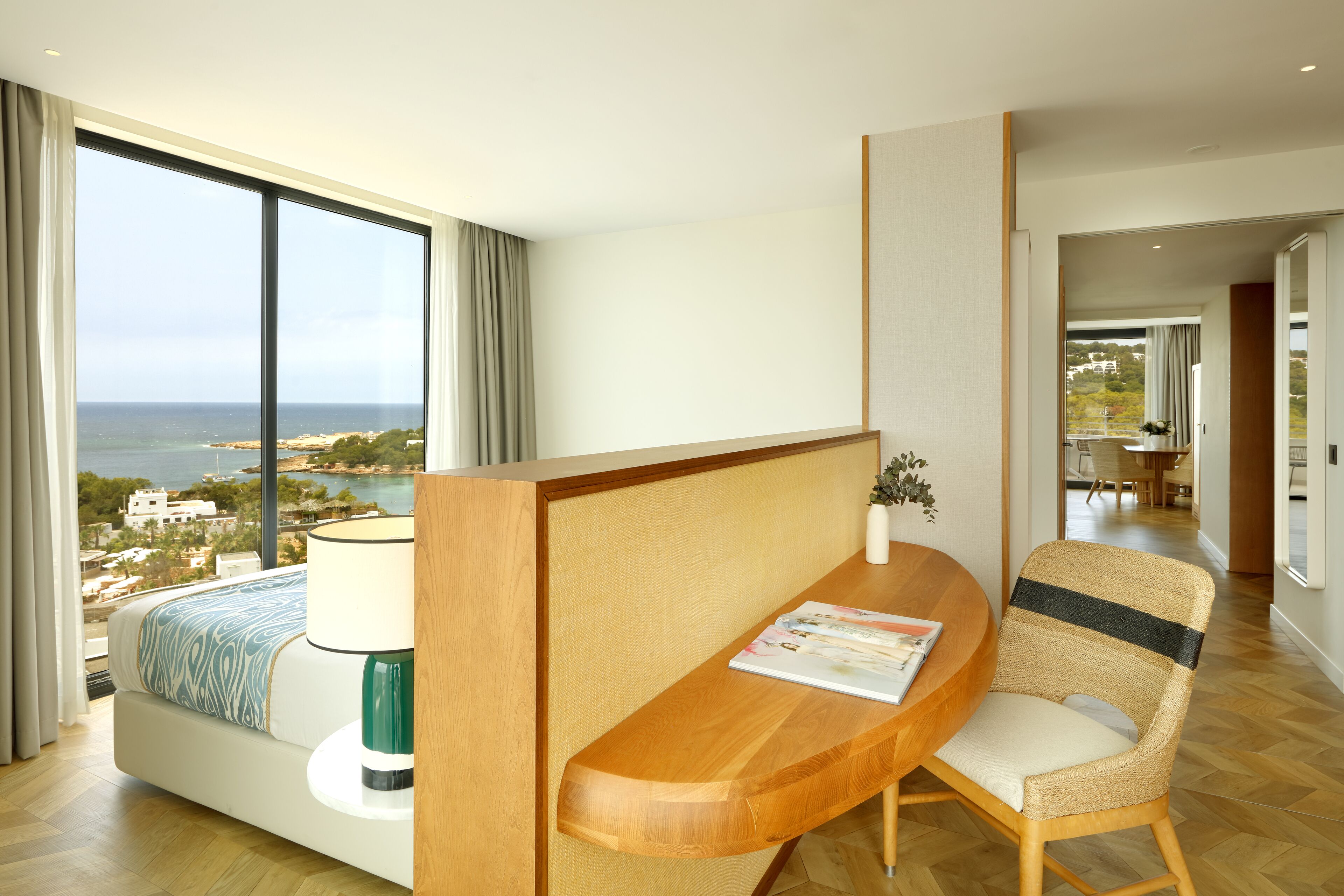 Un cuarto de hotel con un ventanal grande, una cama con manta estampada, un escritorio de madera con lámpara, una silla de mimbre y vista al mar y edificios.