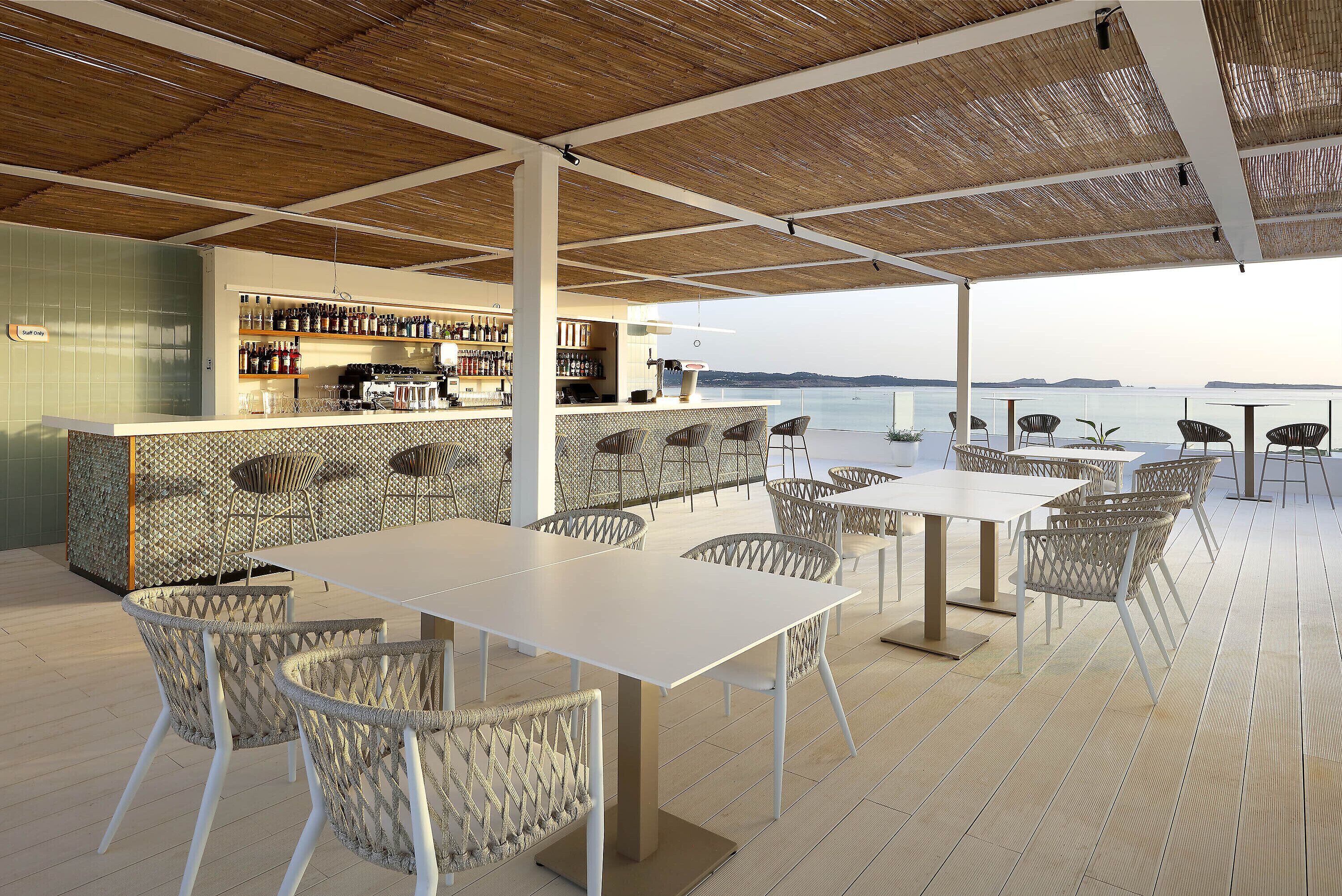 Zona de bar al aire libre con vistas al mar, que incluye una barra, asientos y suelo de baldosas.