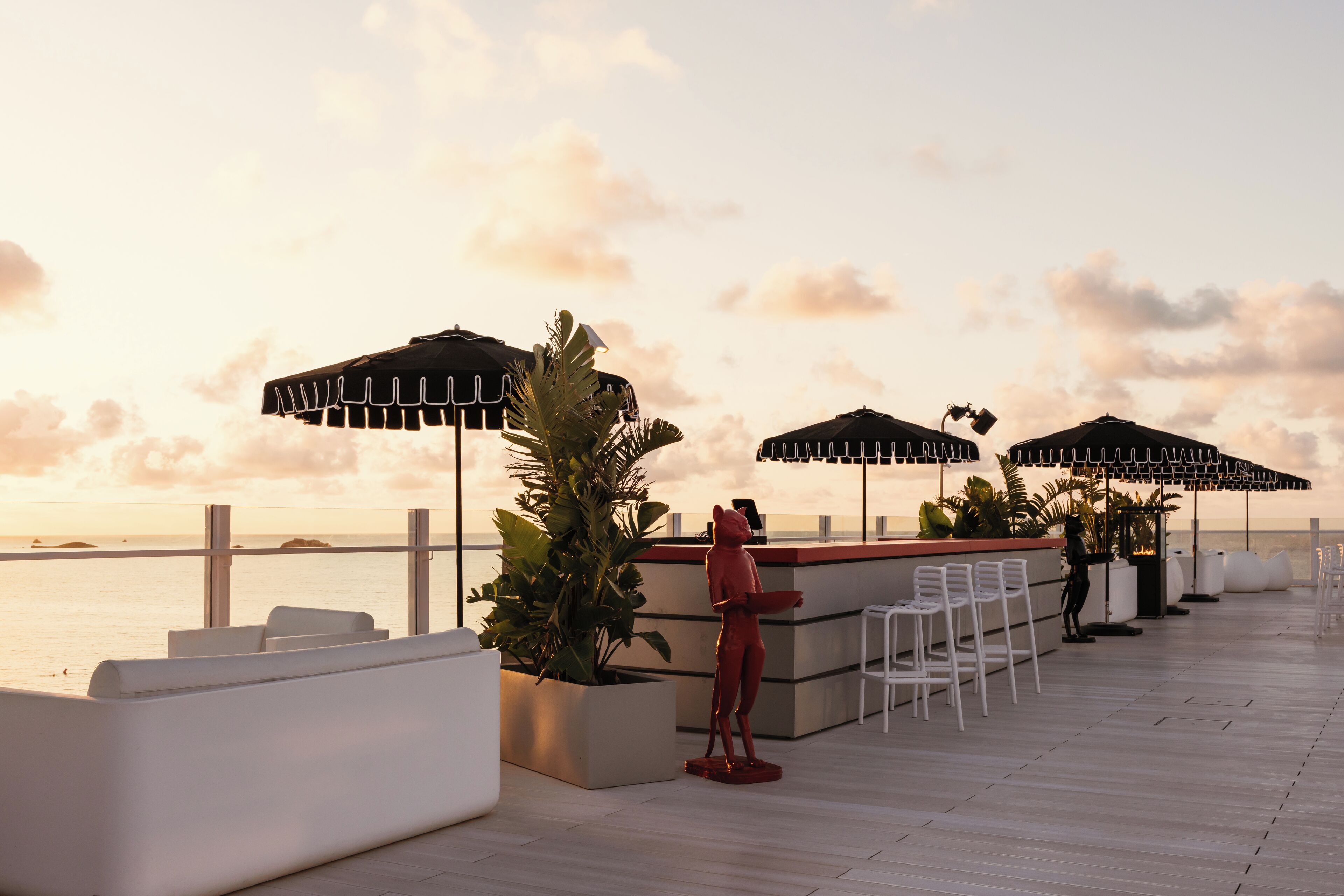 A rooftop bar with outdoor seating, umbrellas, and a view of the ocean at sunset.