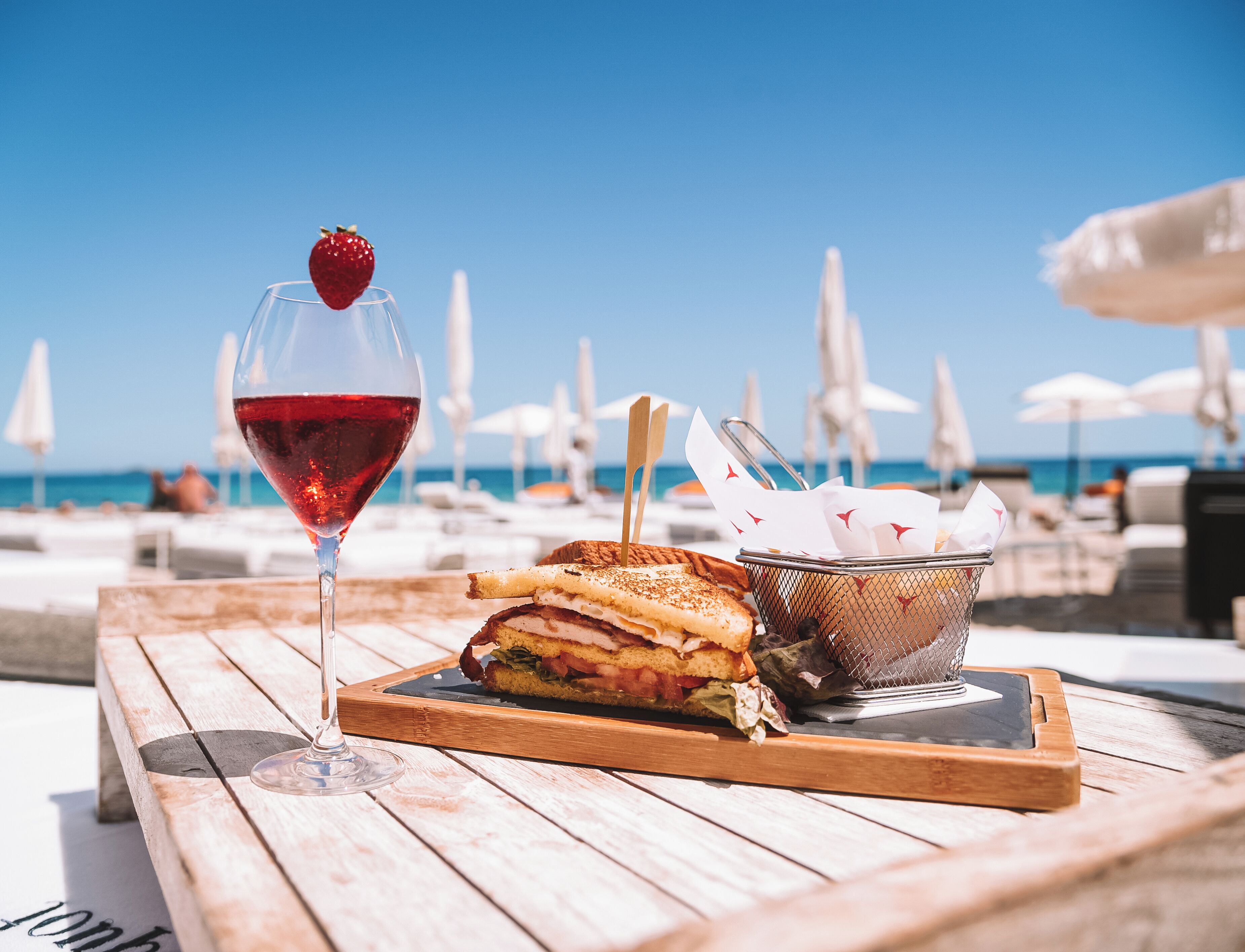 Una mesa de madera con una copa de vino tinto, un sándwich y una cesta de servilletas en un entorno playero.