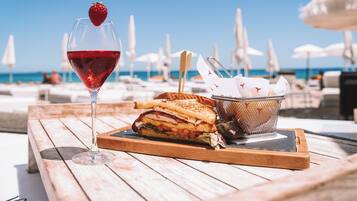 Una mesa de madera con una copa de vino tinto, un sándwich y una cesta de servilletas en un entorno playero.