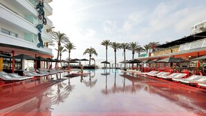 A pool area with lounge chairs and umbrellas, surrounded by palm trees and a building with a modern design. - The Unexpected Ibiza Hotel - Ushuaïa Club entrance included (Sant Josep de sa Talaia)