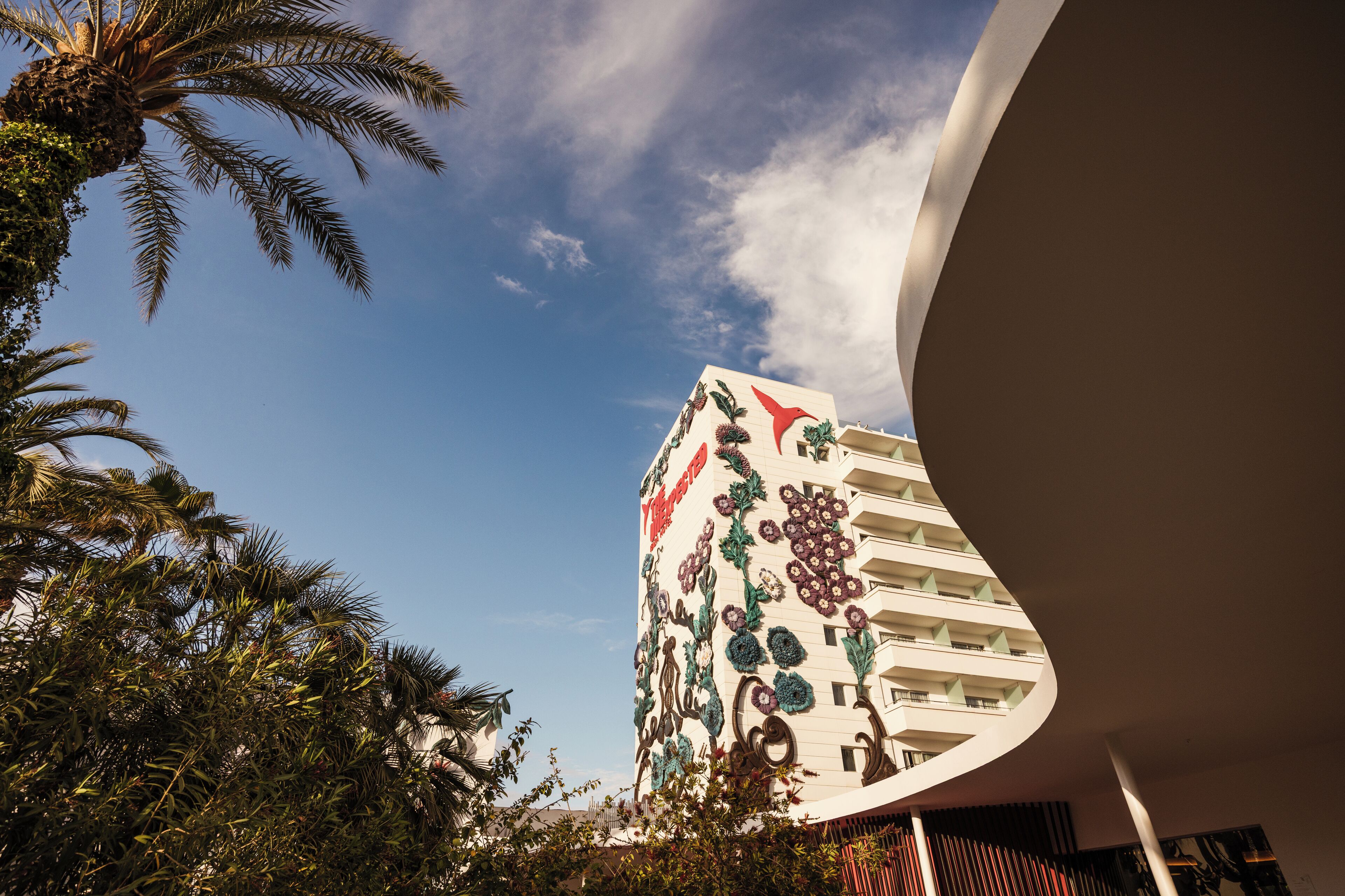 Un hotel con fachada curva, un mural floral y palmeras en primer plano.