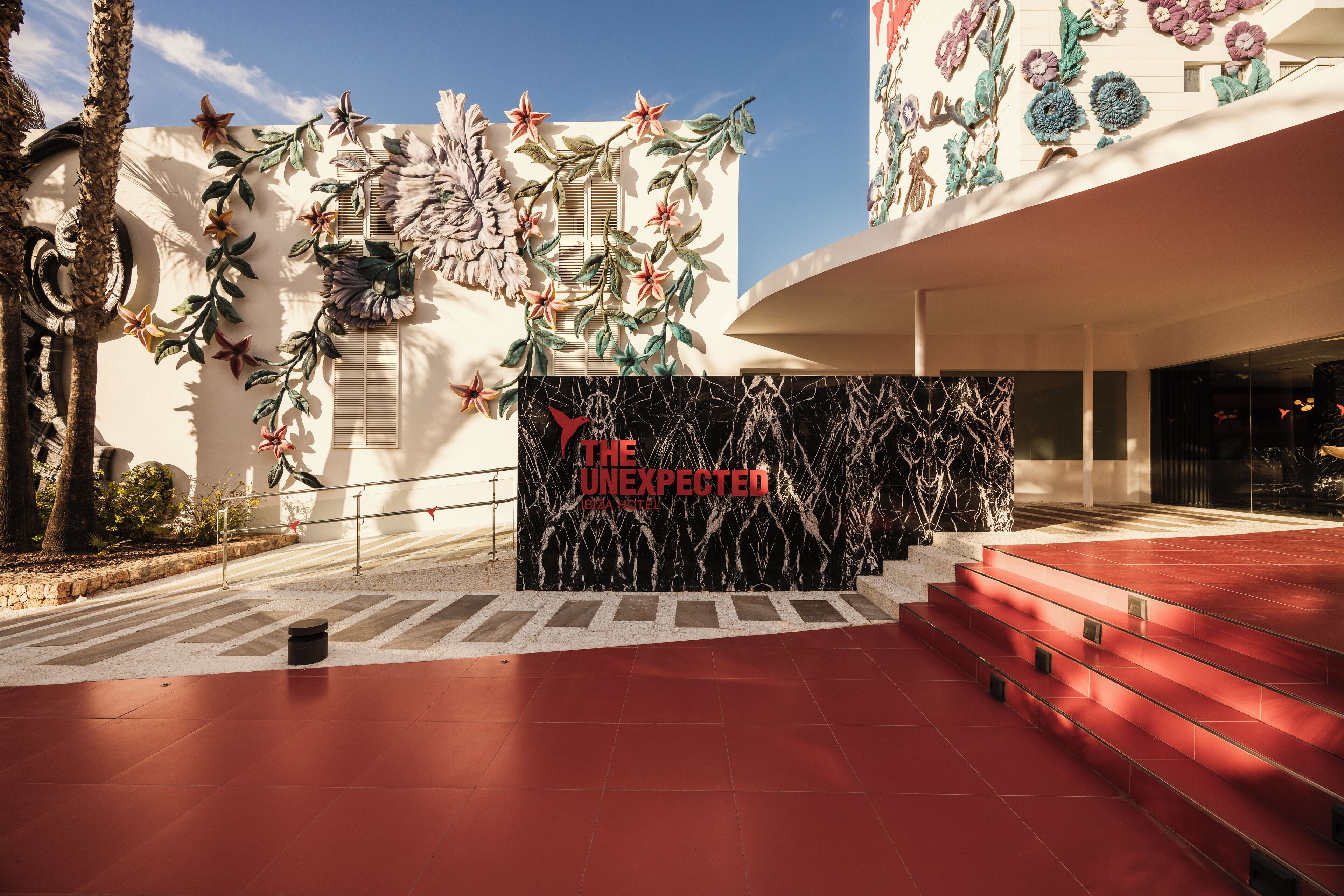 A building entrance with a red tiled floor, a black marble wall with 'THE UNEXPECTED' text, and a decorative floral mural.