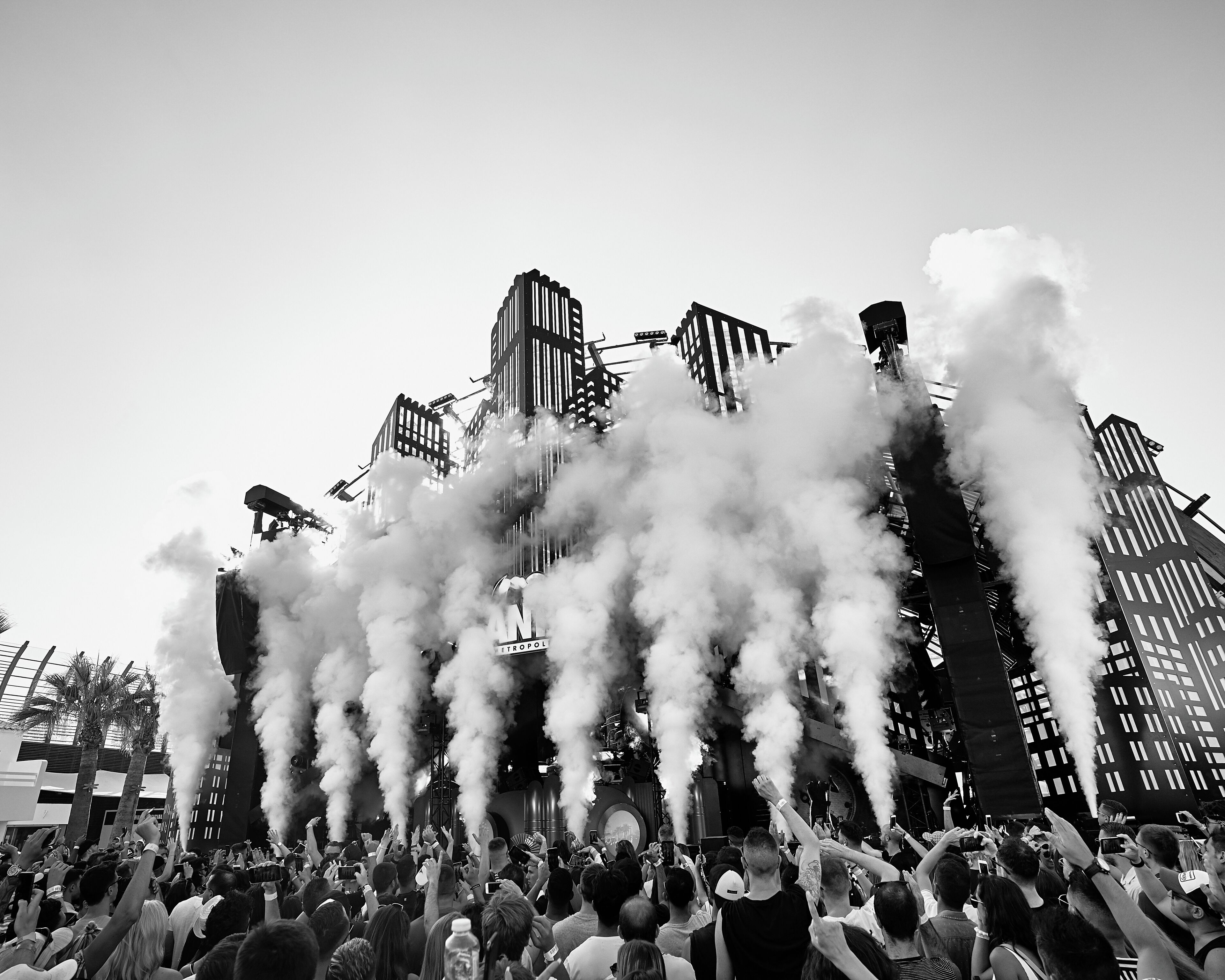 Un concierto con una multitud frente a un escenario con efectos de humo, rodeado de edificios altos.