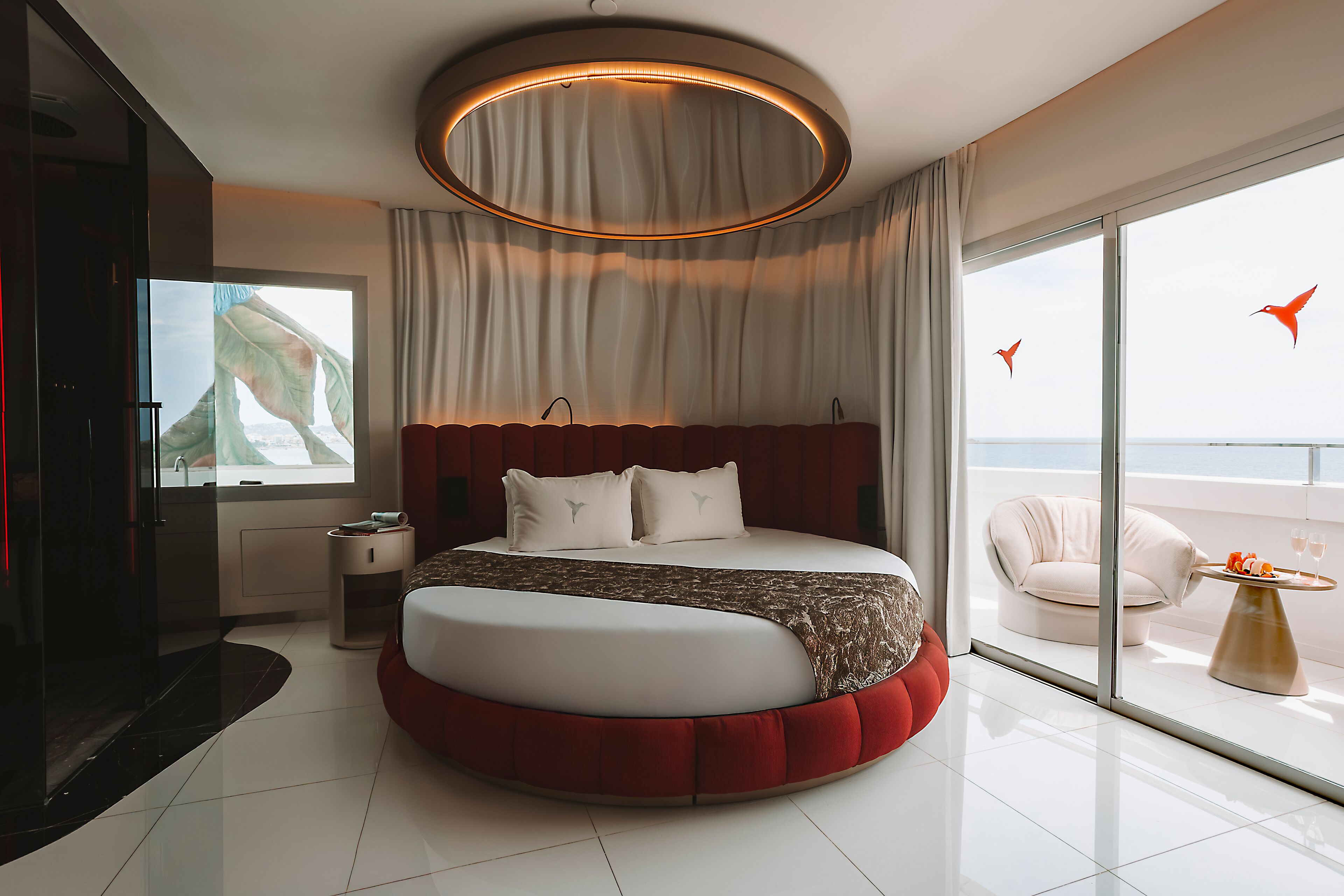 Un dormitorio moderno con una cama redonda, televisión, balcón con vistas y una lámpara de techo circular.