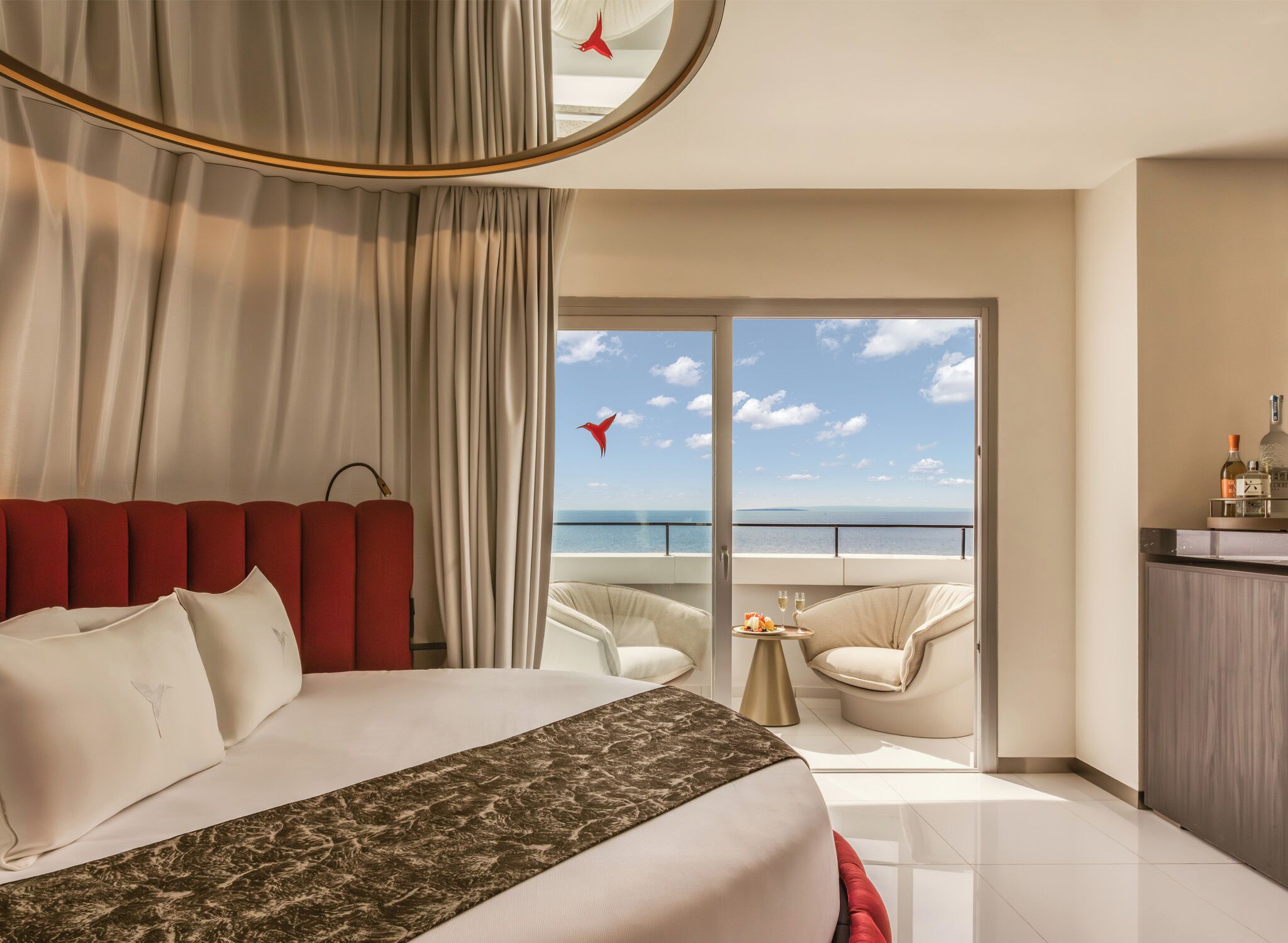 A hotel room with a bed, a balcony with a view of the sea, a dining area, and a minibar.